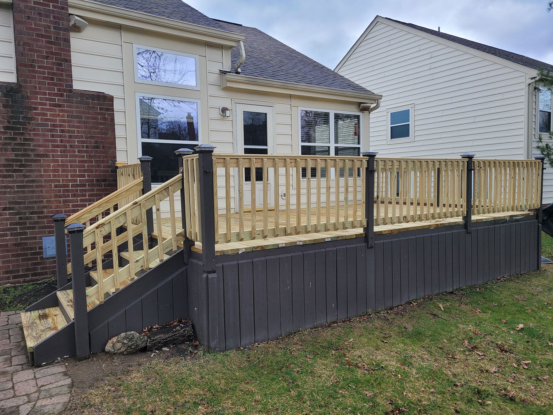 Wooden deck with railing attached to a beige house, with dark grey base and surrounding grass.