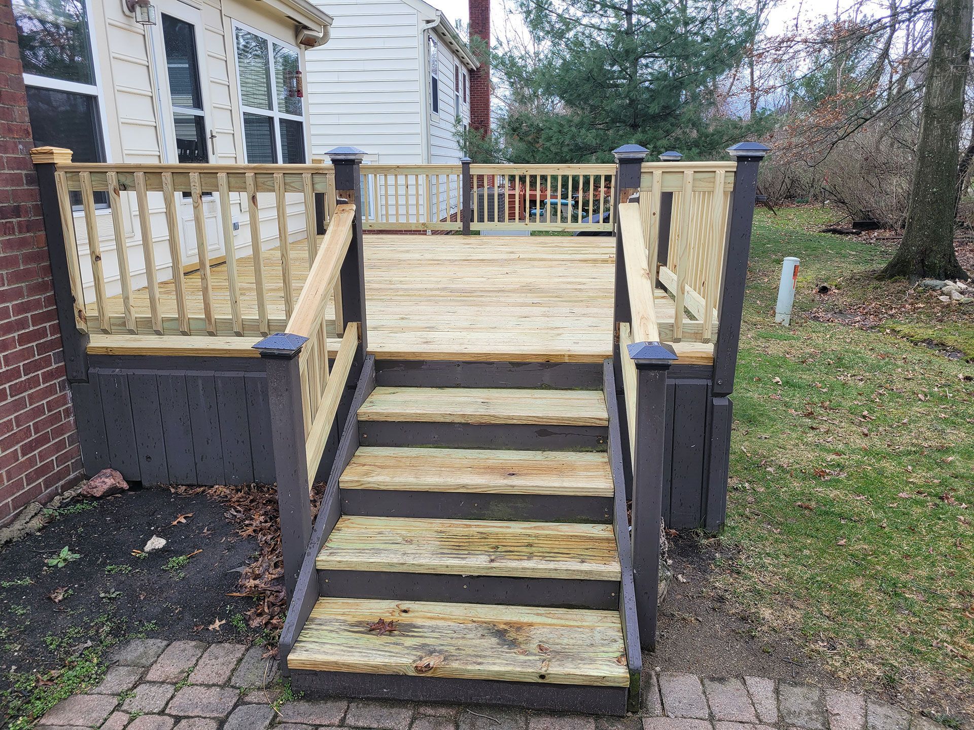 Wooden deck with stairs, gray and light wood tones. Brick patio in foreground, green lawn in background.
