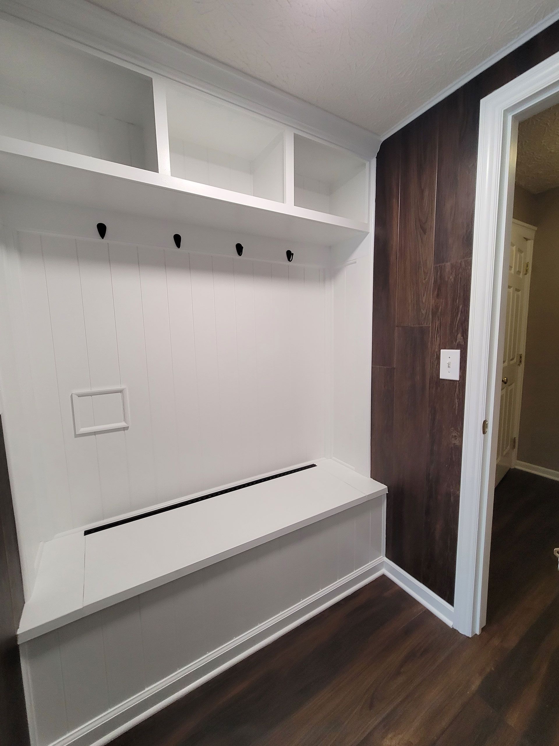 Built-in white entryway bench with shelves and coat hooks. Dark wood paneling and a doorway are visible.
