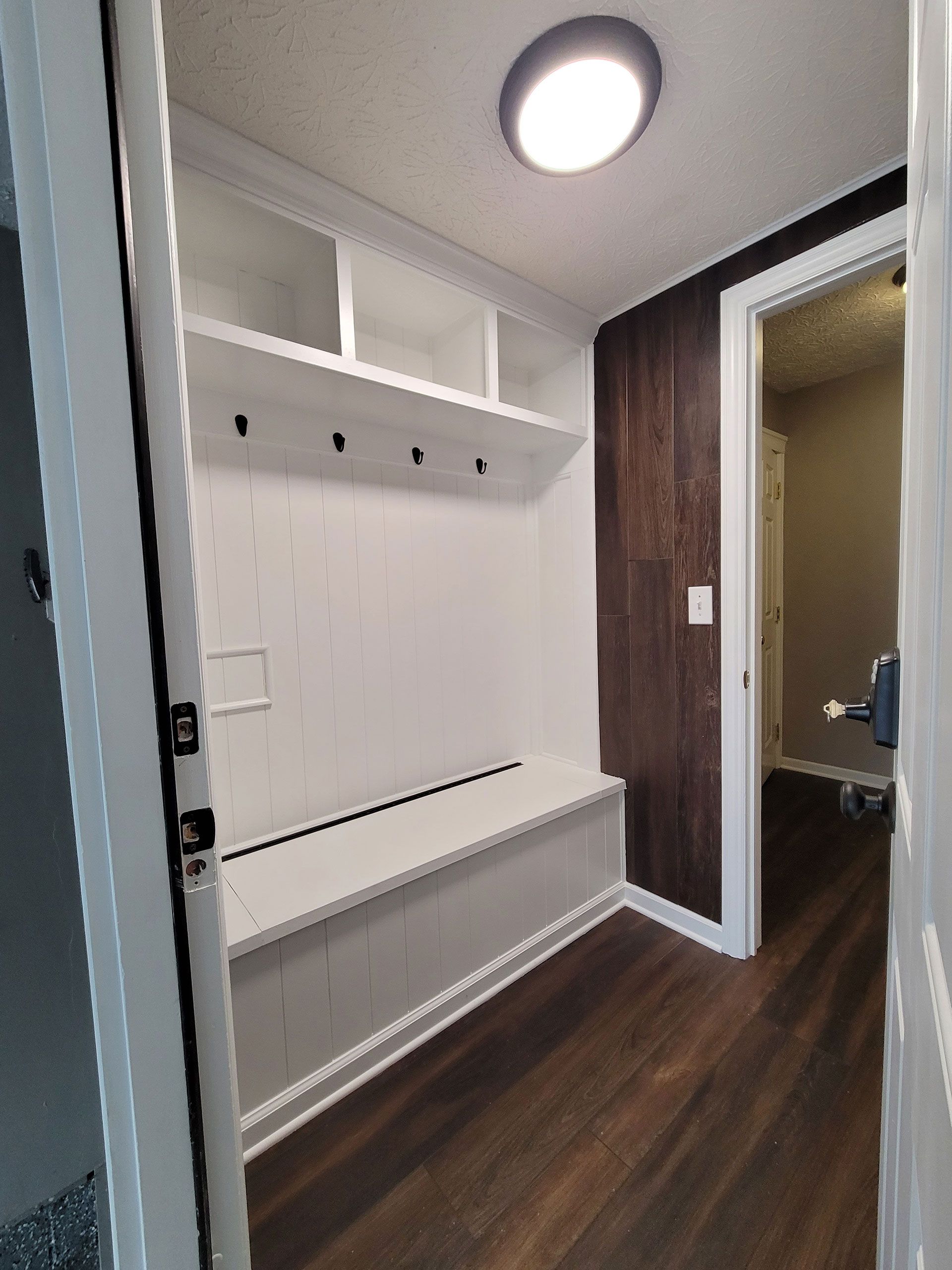 Mudroom with built-in bench and shelves; dark wood wall, light wood floor, white trim, hooks, and ceiling light.