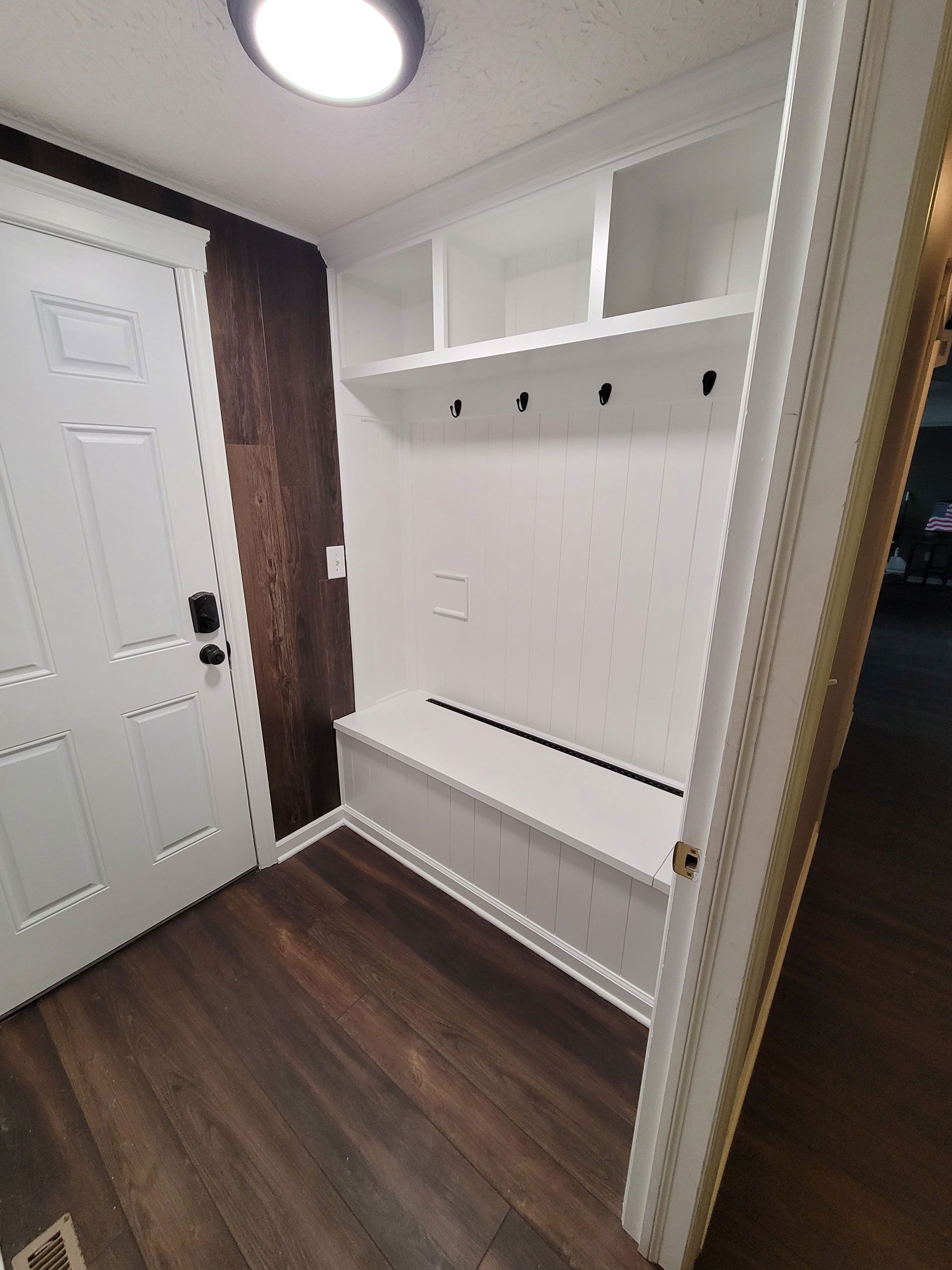 White built-in storage bench with hooks, shelves, and cabinets in entryway with dark wood flooring and white door.