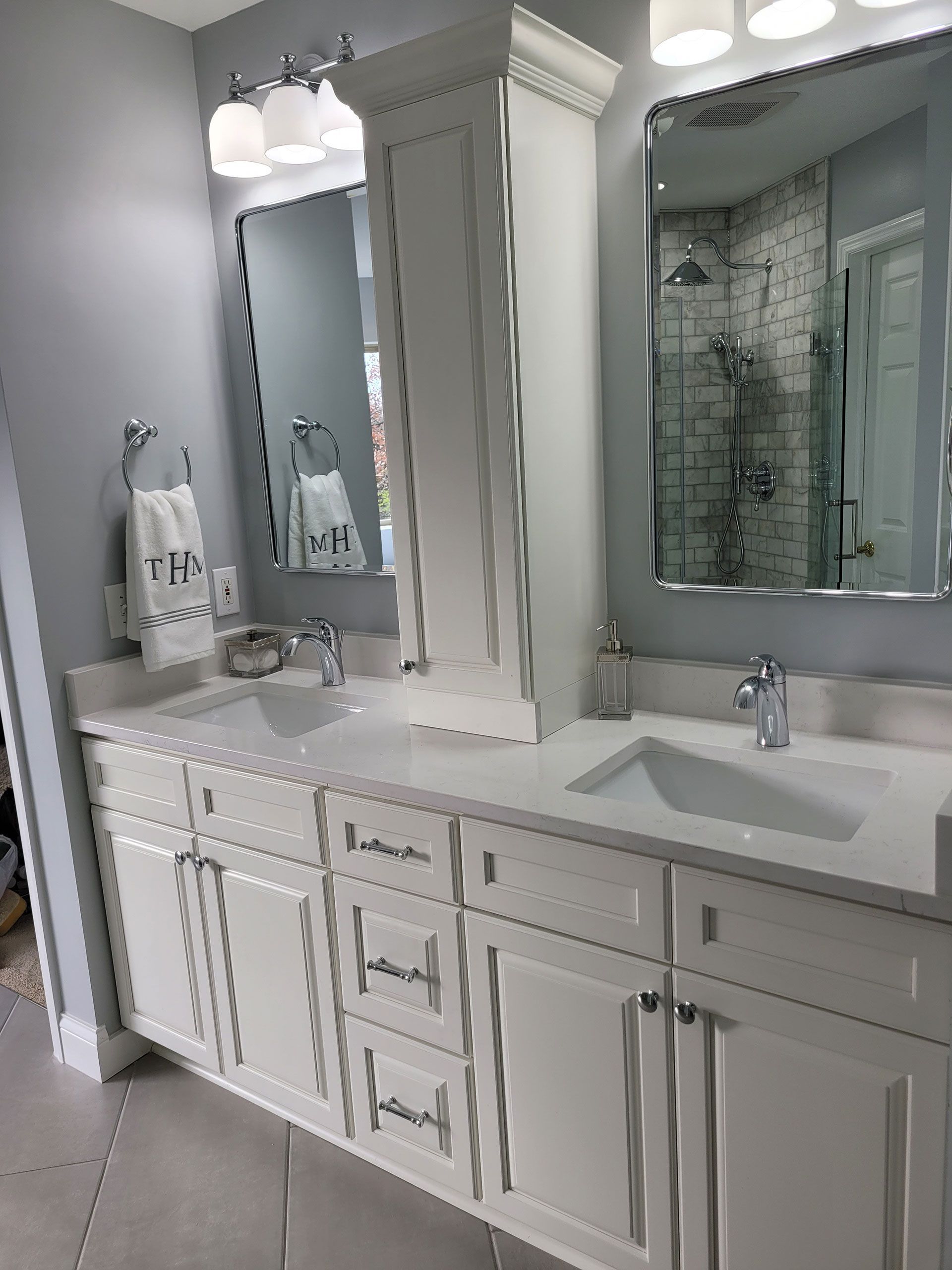 Bathroom with white cabinetry, light countertops, and two sinks. Gray walls and mirrors reflect the shower.