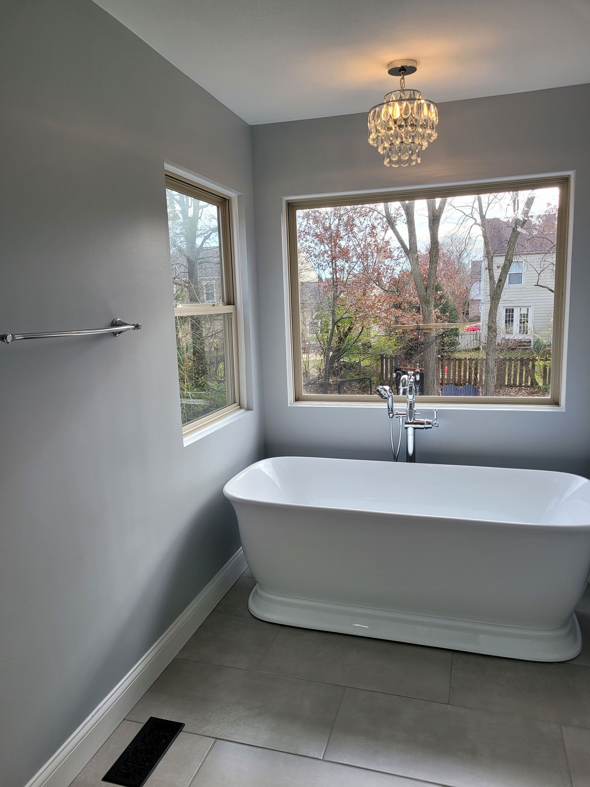 Bathroom with a white freestanding bathtub beneath a window with outdoor trees visible. Gray walls, chandelier.