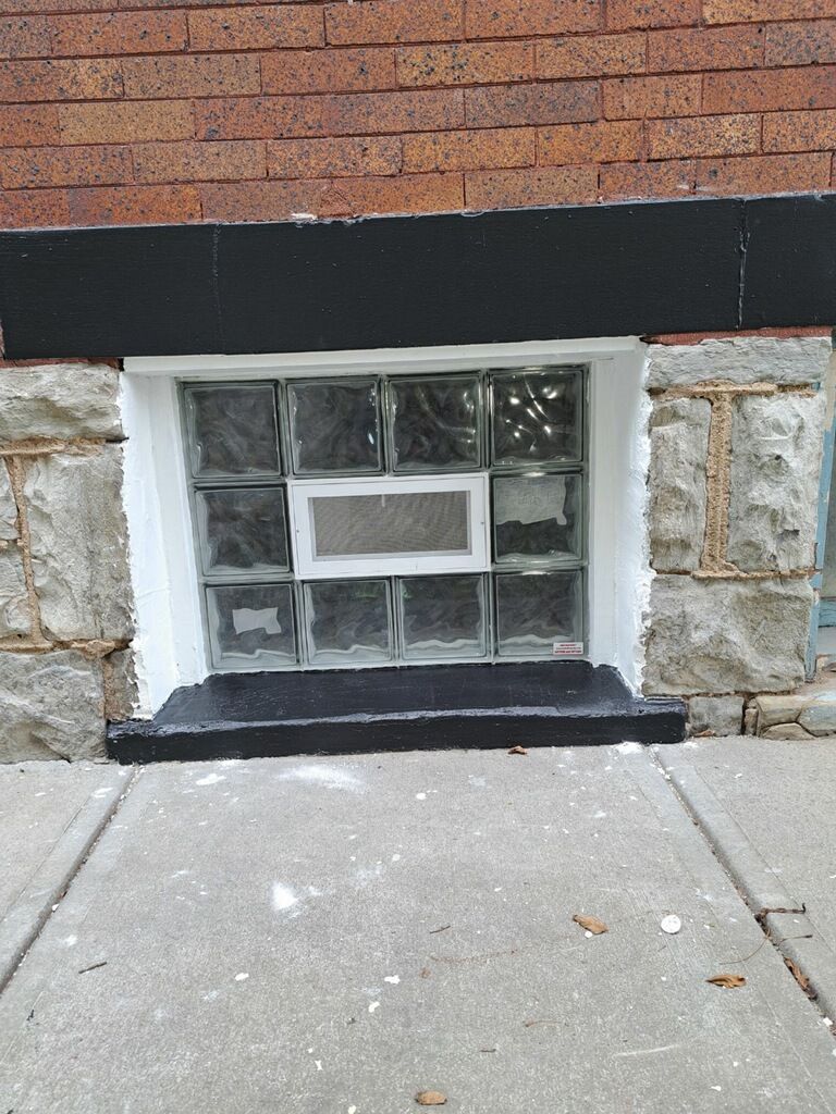 Glass block window in a brick building's exterior wall, framed by stone and topped with a black painted border.
