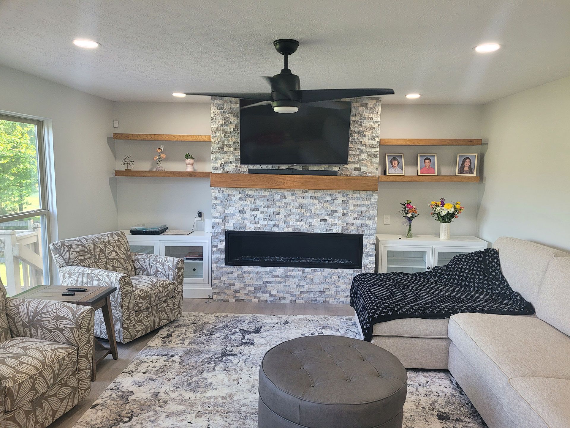 Living room with gray stone fireplace, TV, and shelves, neutral-toned furniture, and a round ottoman on a patterned rug.
