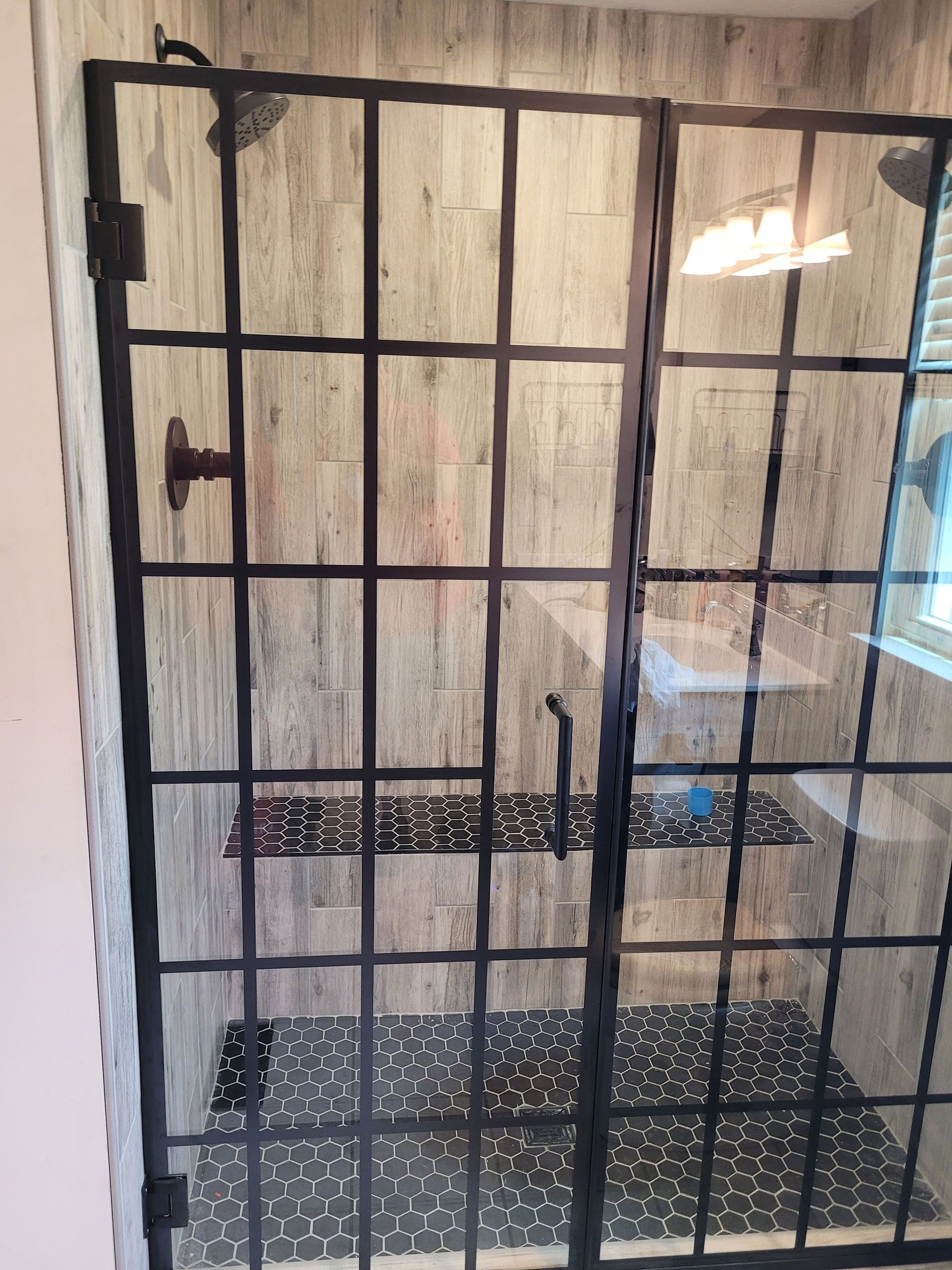 Shower enclosure with black grid frame and glass panels, light wood-look tile, dark mosaic floor.
