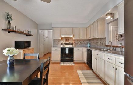 Kitchen with black appliances, light cabinets, wooden floor, dining table, and open to a hallway.