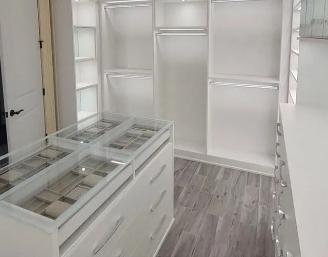 White walk-in closet with shelves, drawers, and hanging rods.  Gray wood-look flooring. Glass-top island in foreground.
