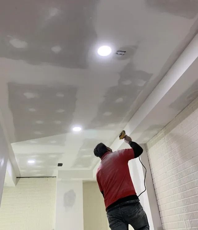 Person sanding a ceiling, drywall patches visible, in a room with recessed lights and white walls.