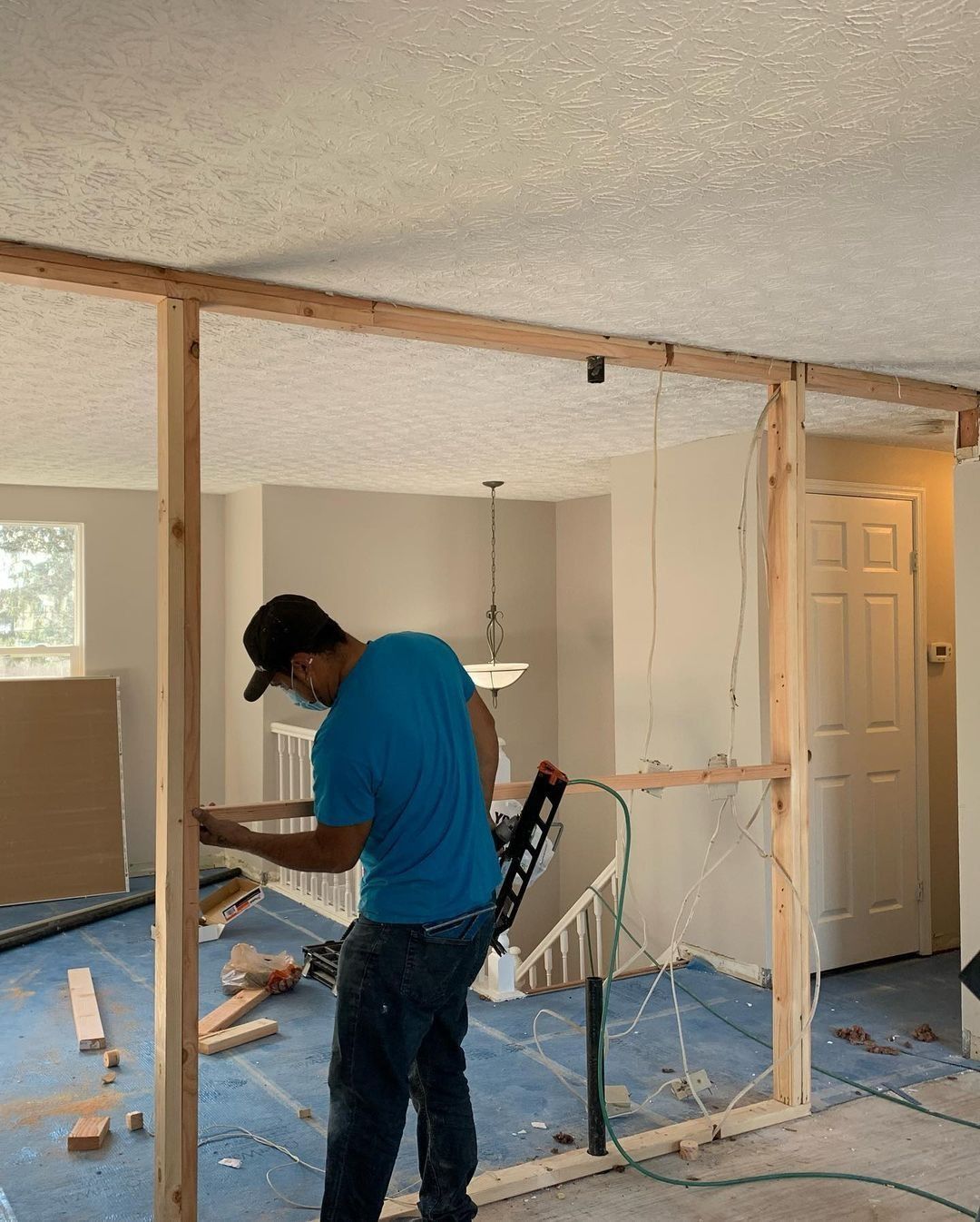 A person constructing a wooden wall frame indoors. The person is wearing a blue shirt and jeans.