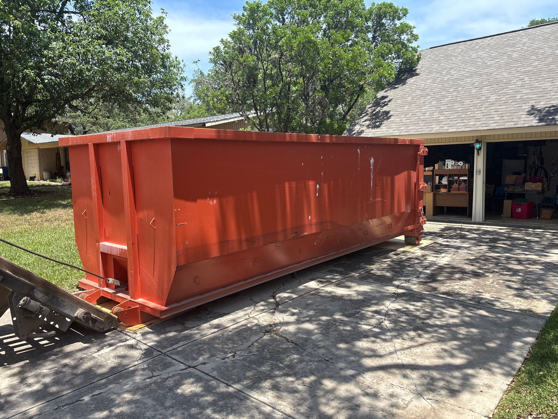 Large orange dumpster in a residential driveway, partially obscuring a garage and house under a blue sky.