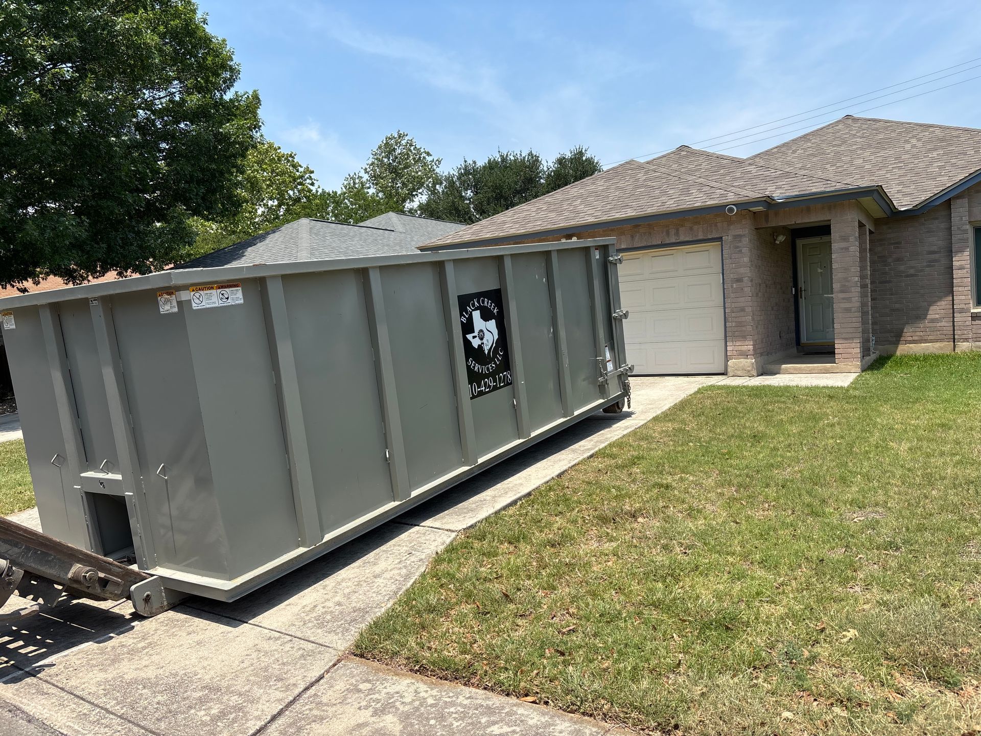 Gray dumpster parked in front of a brick house on a sunny day.