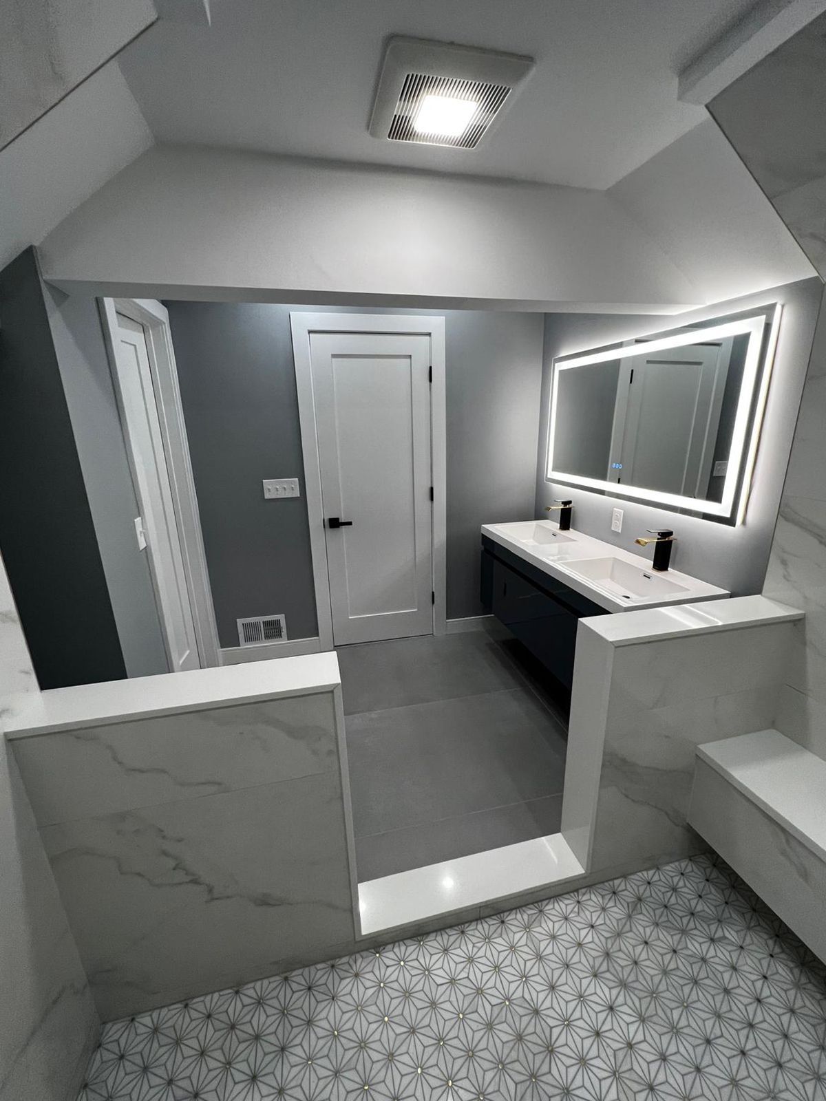 Modern bathroom with a large lit mirror, gray walls, dark vanity, and patterned floor tile.