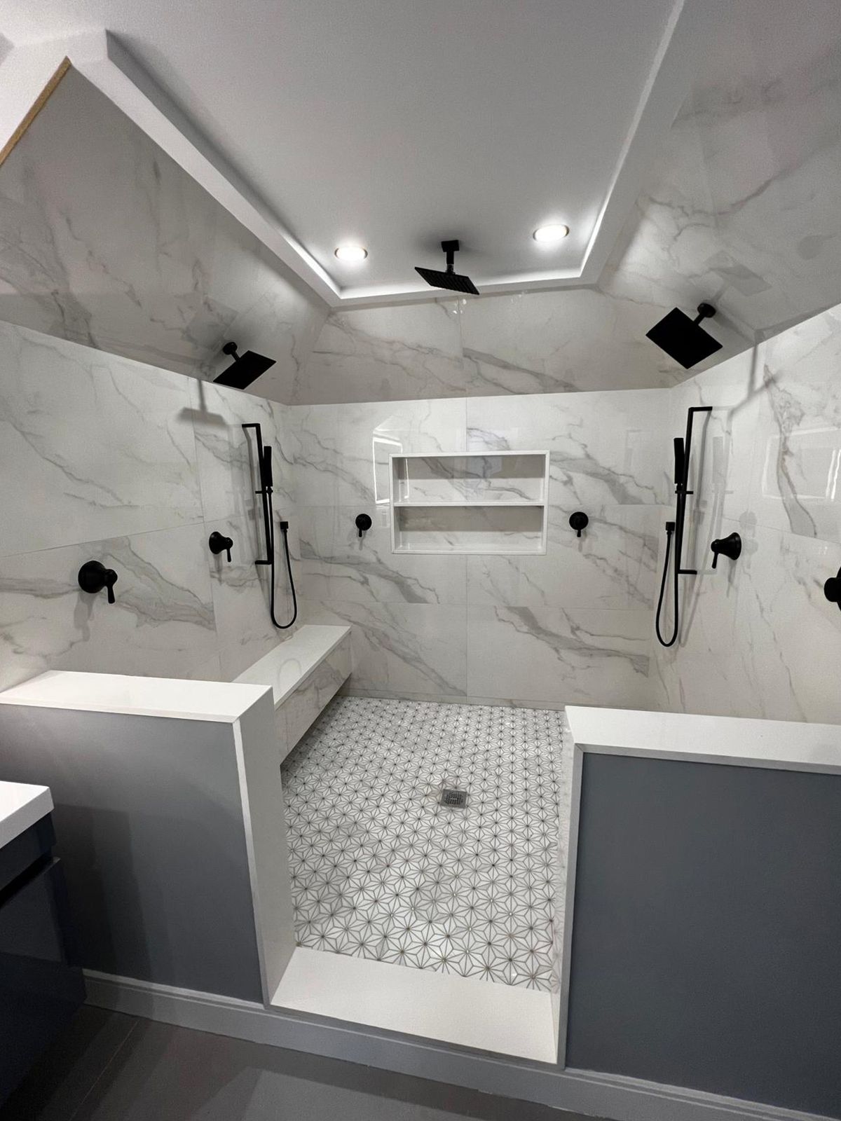 Spacious, tiled shower with multiple showerheads. Features a bench and inset shelves. Black fixtures against white and gray tiles.
