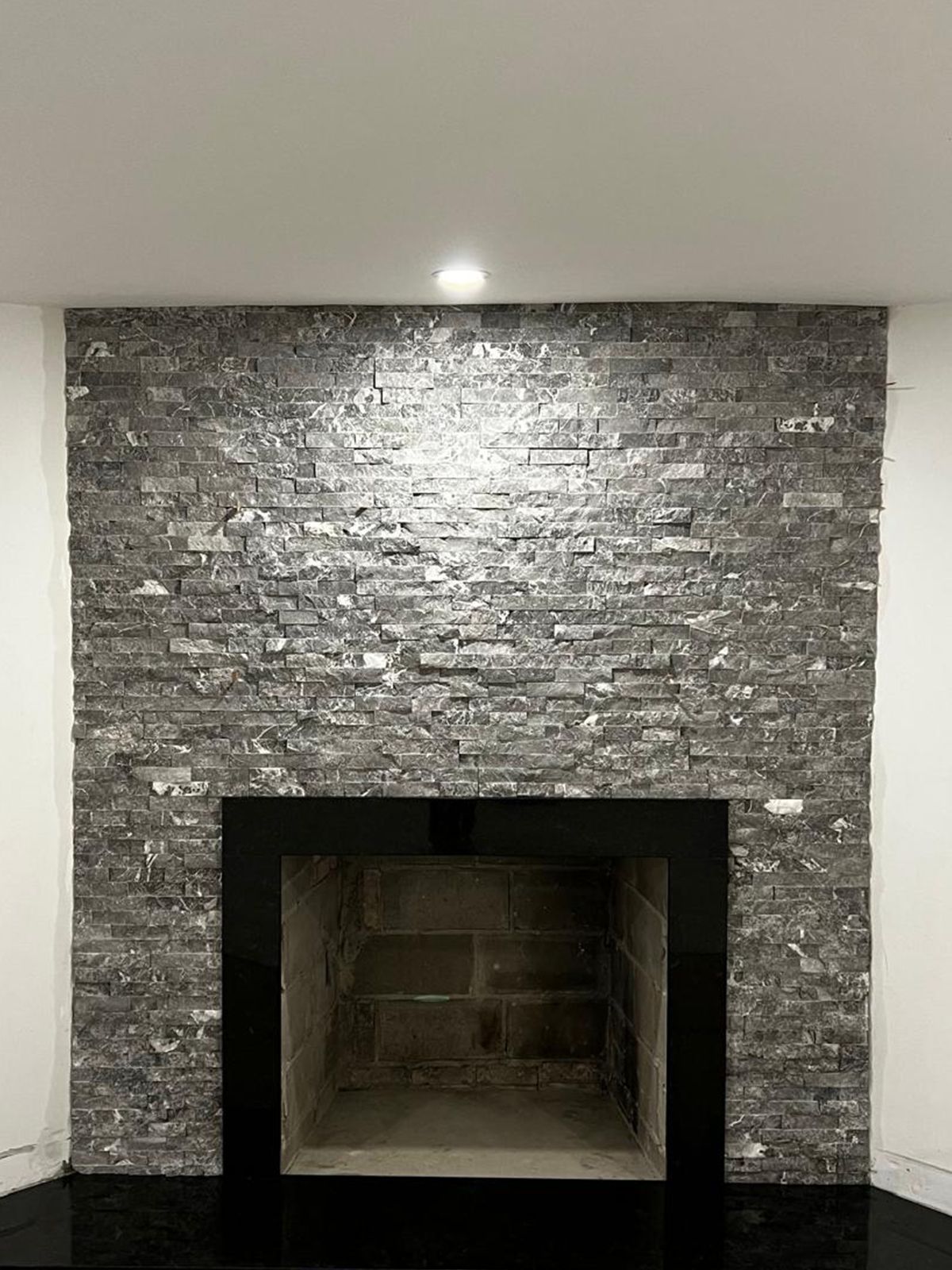 Stone-clad fireplace with black trim and a dark hearth, set against white walls, lit by a centered overhead light.