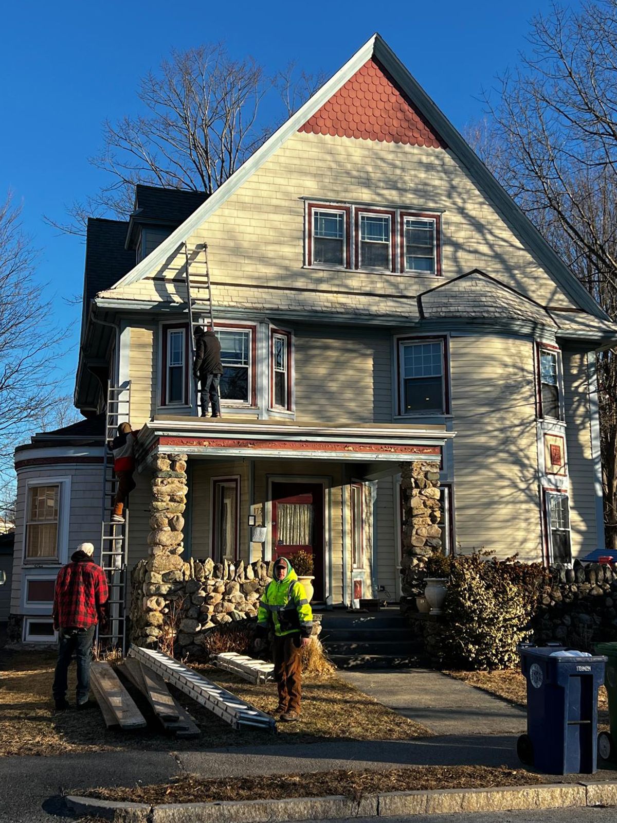 Workers on ladders, renovating a light-colored house with stone pillars and a red roof peak.