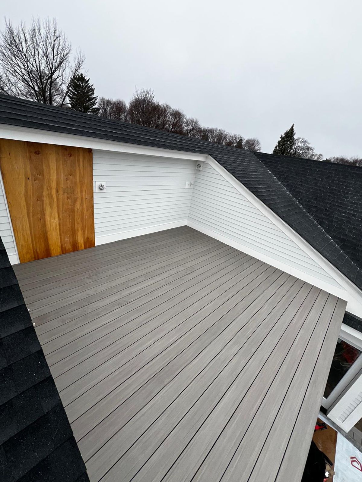 Gray composite deck with white siding and wooden door. Black shingled roof overhead.