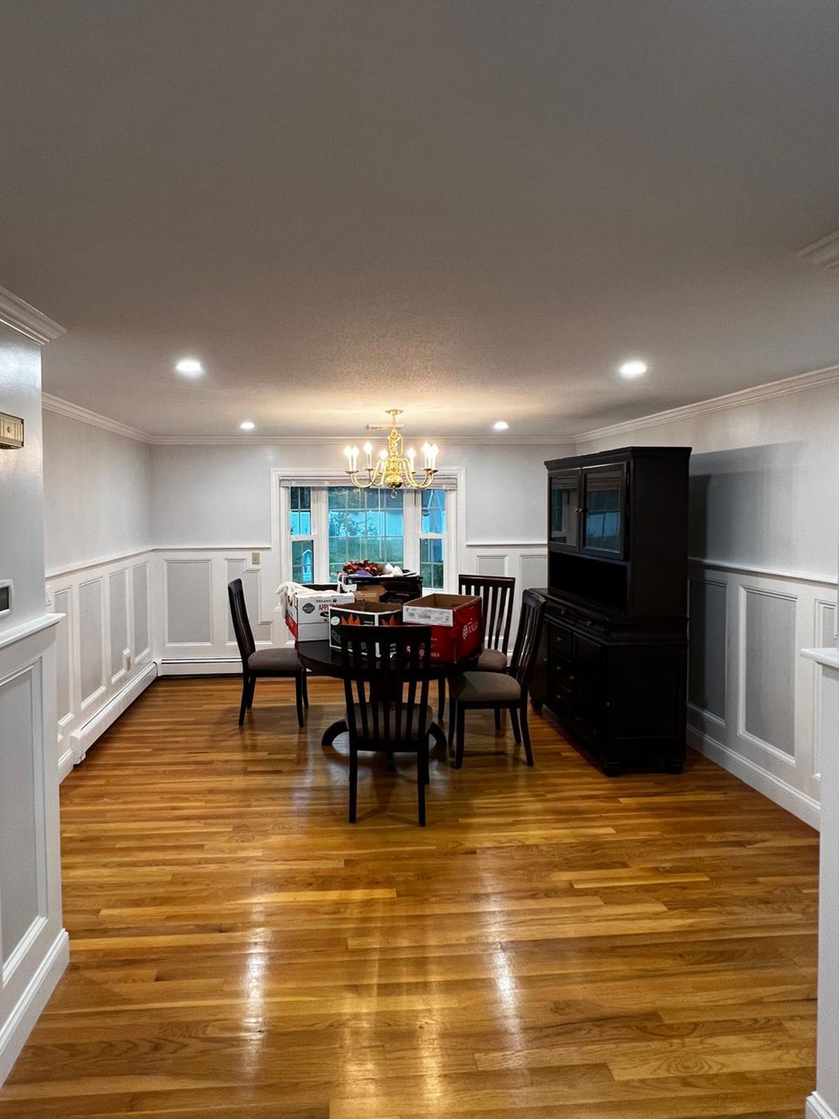 Dining room with hardwood floor, wainscoting, table, chairs, and dark cabinet. Bright lighting from recessed lights and chandelier.
