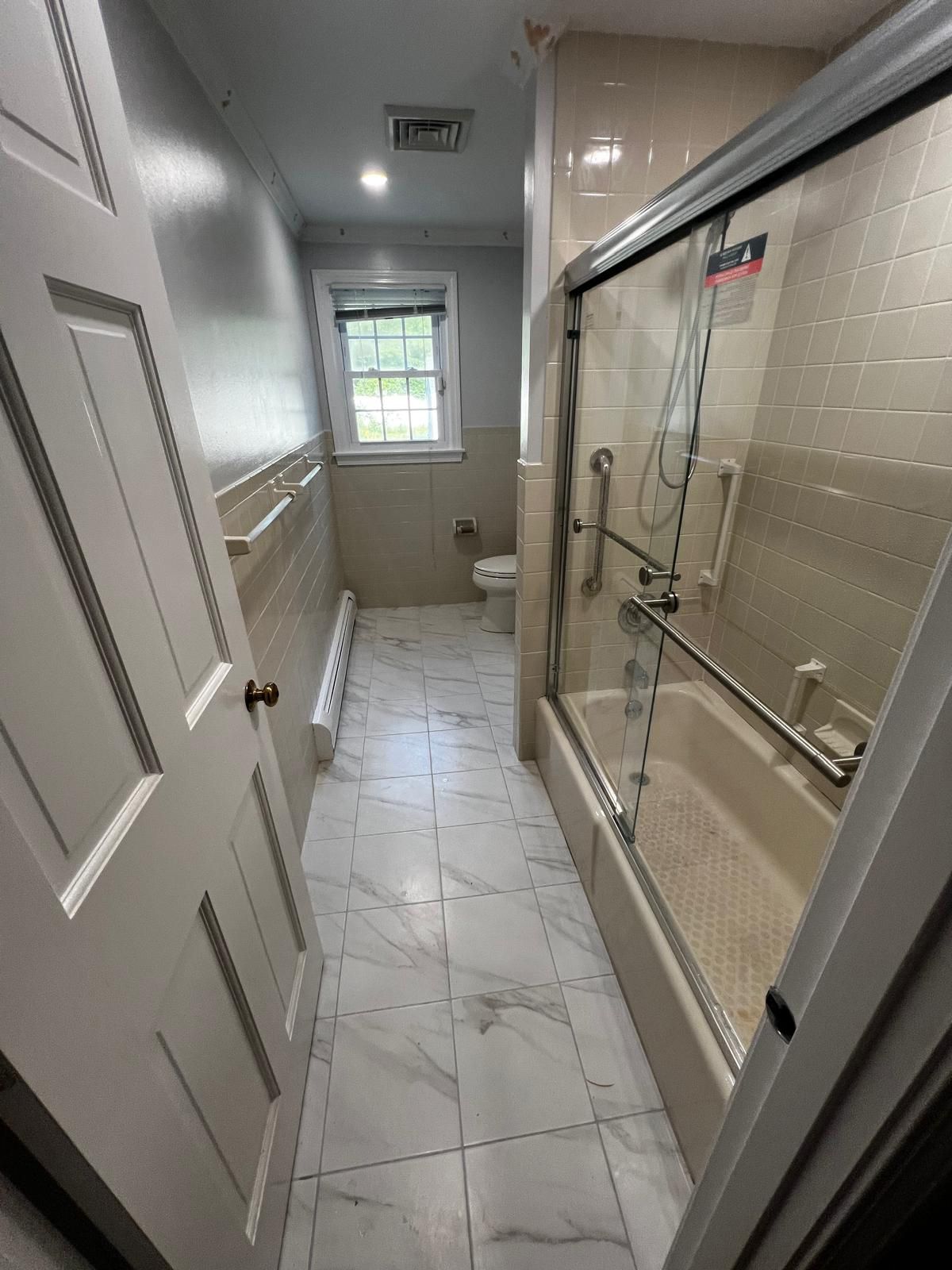 Bathroom with white tile floor, tub with sliding glass doors, toilet, and window.