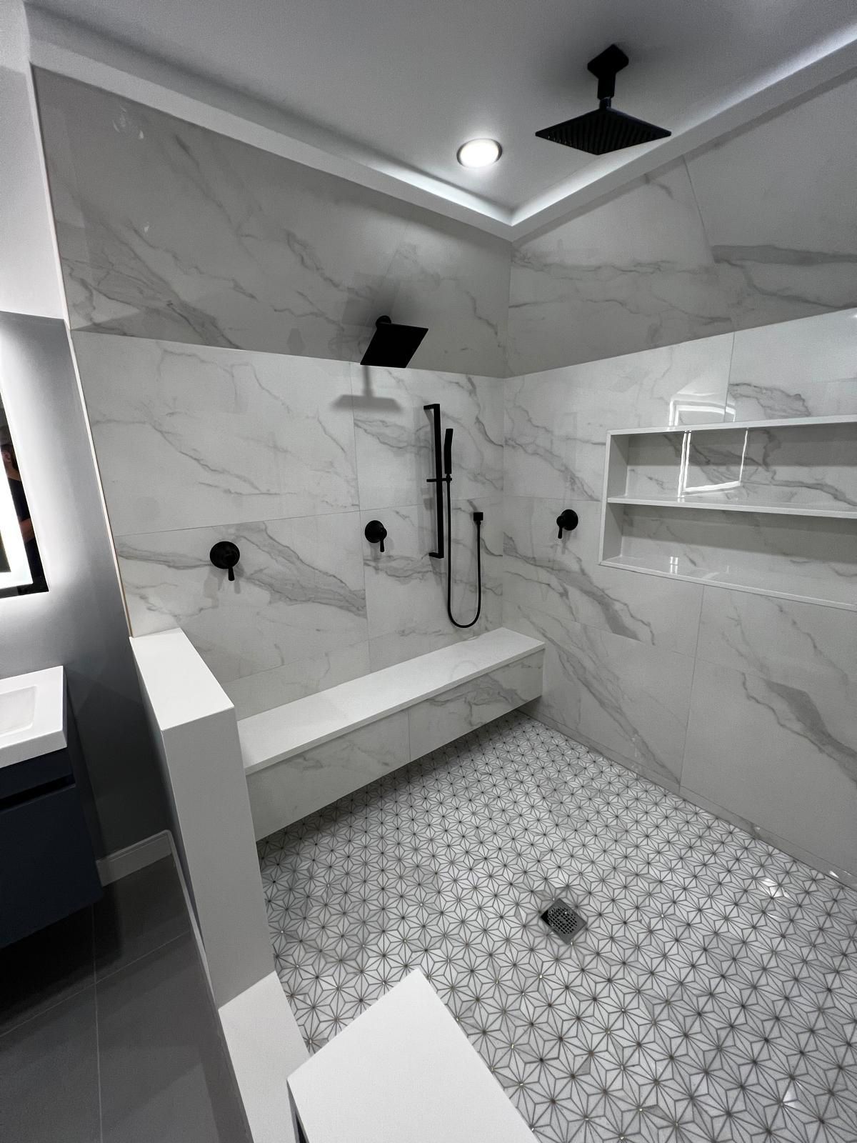 Modern shower with large marble-look tiles, black fixtures, built-in bench, and patterned floor tile.