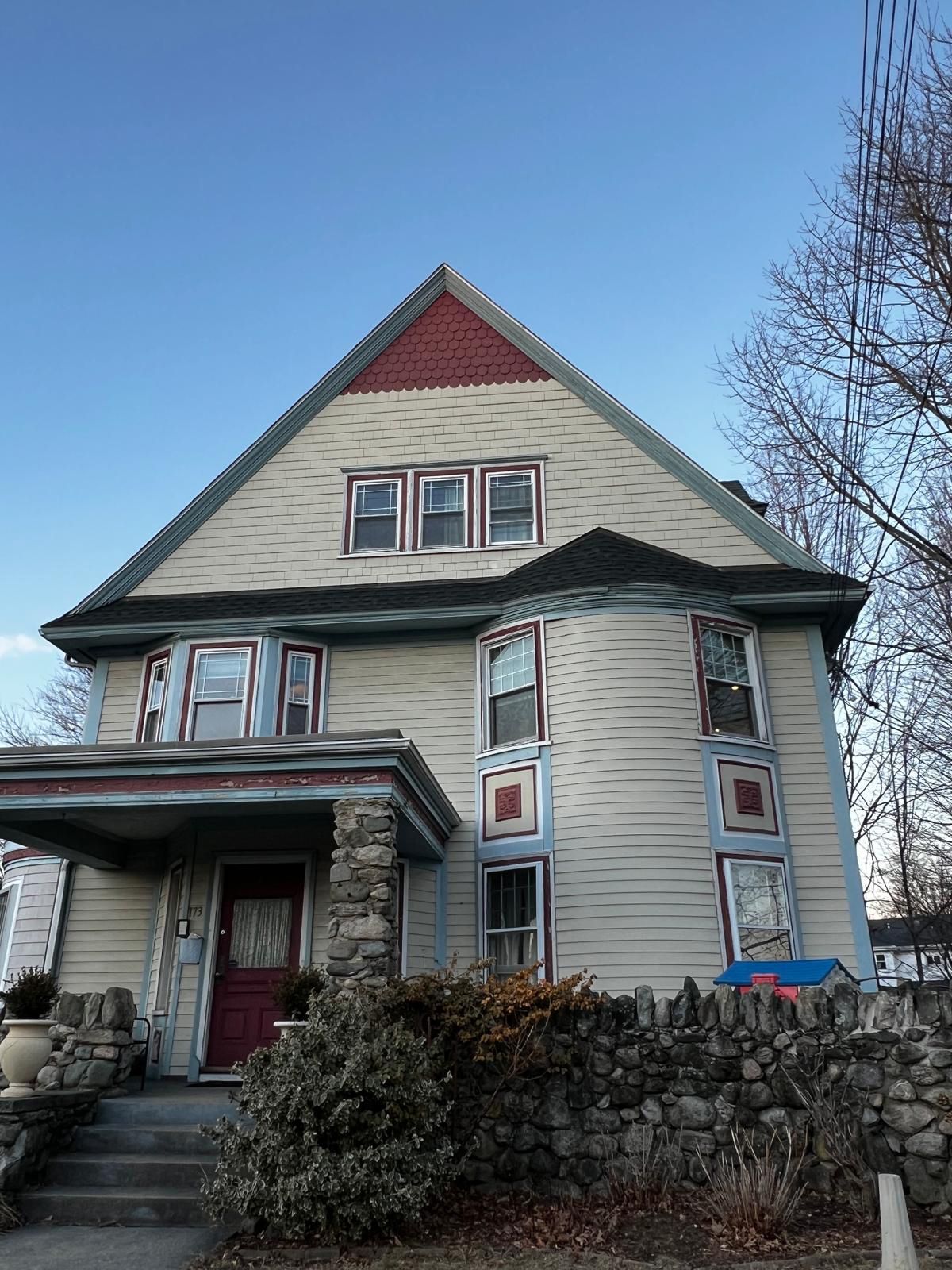 Two-story house with a large triangular roof and bay windows; stone wall and porch.