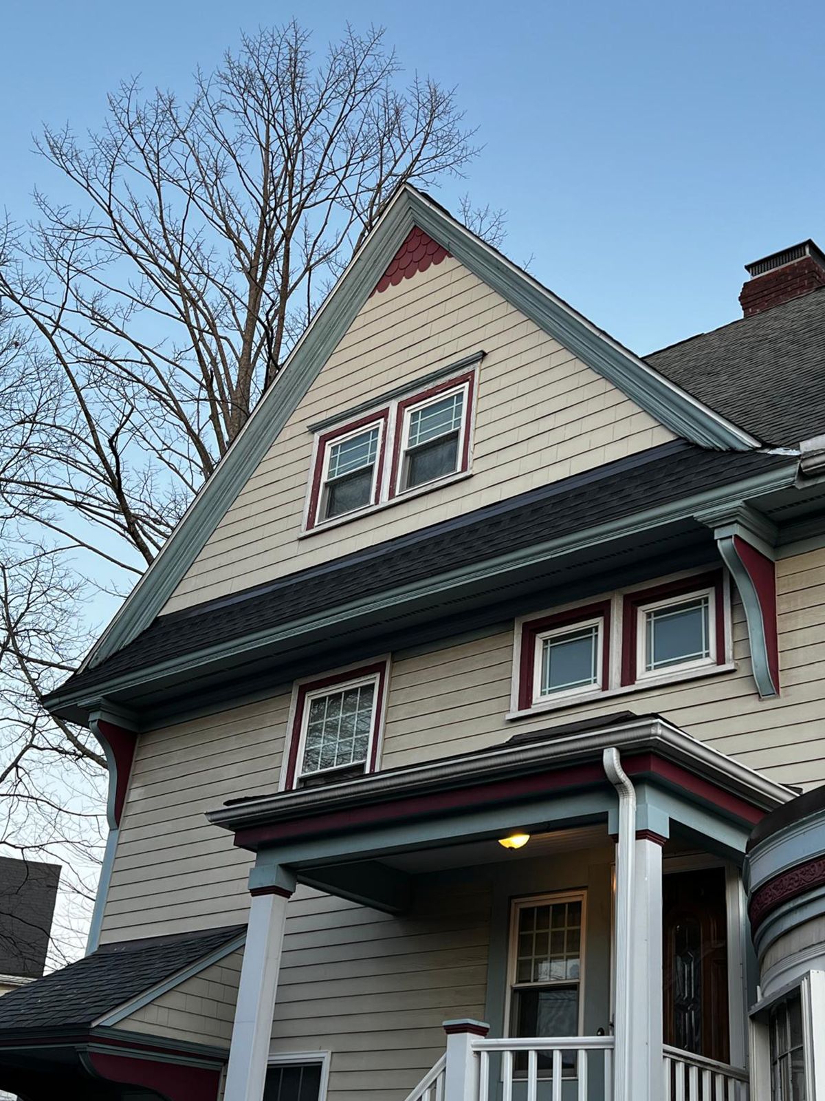 Tan house with gabled roof, red and blue trim, multiple windows, and a bare tree against a blue sky.