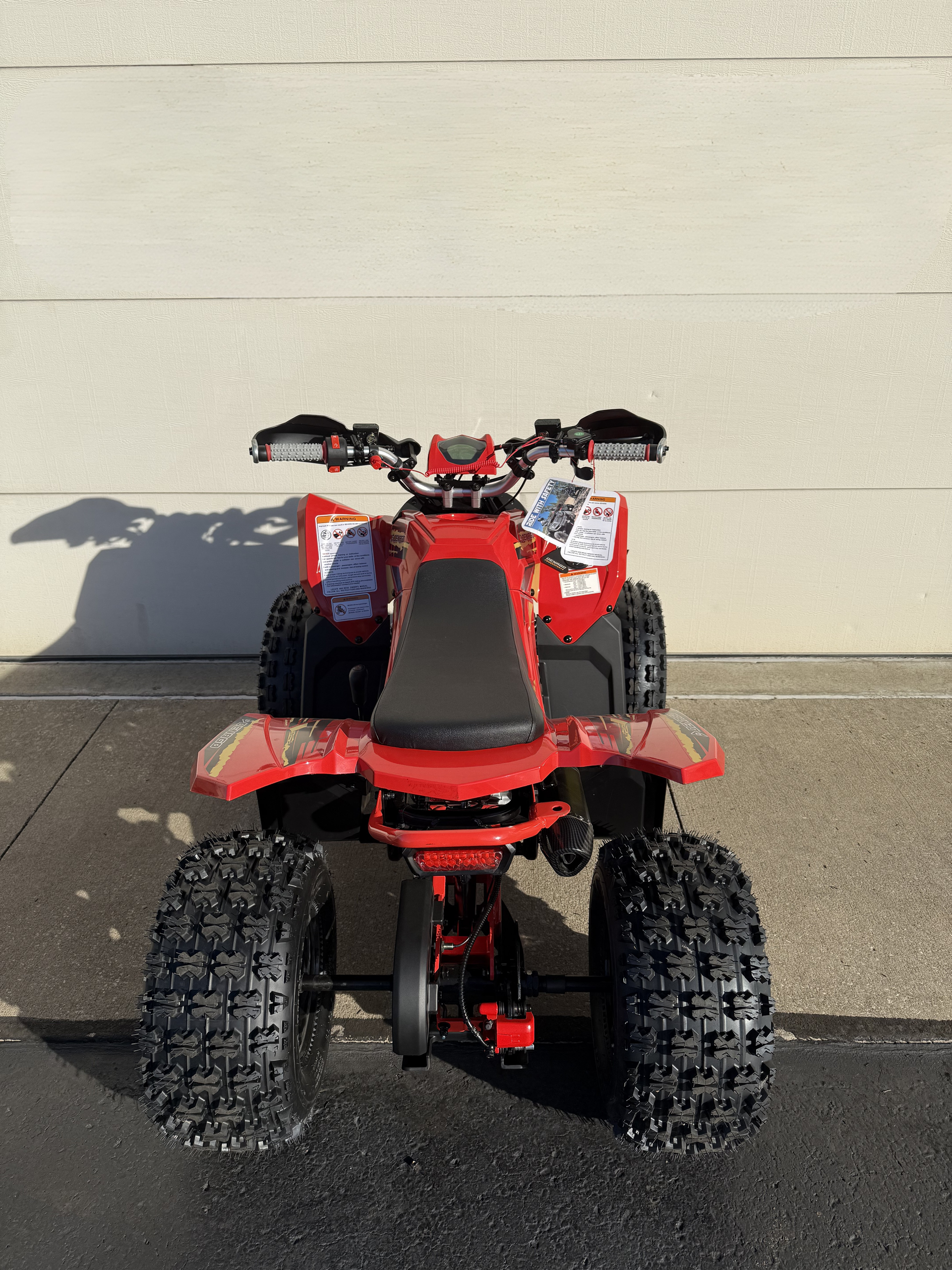 A red ATV sits parked on concrete in front of a white wall, viewed from directly behind.