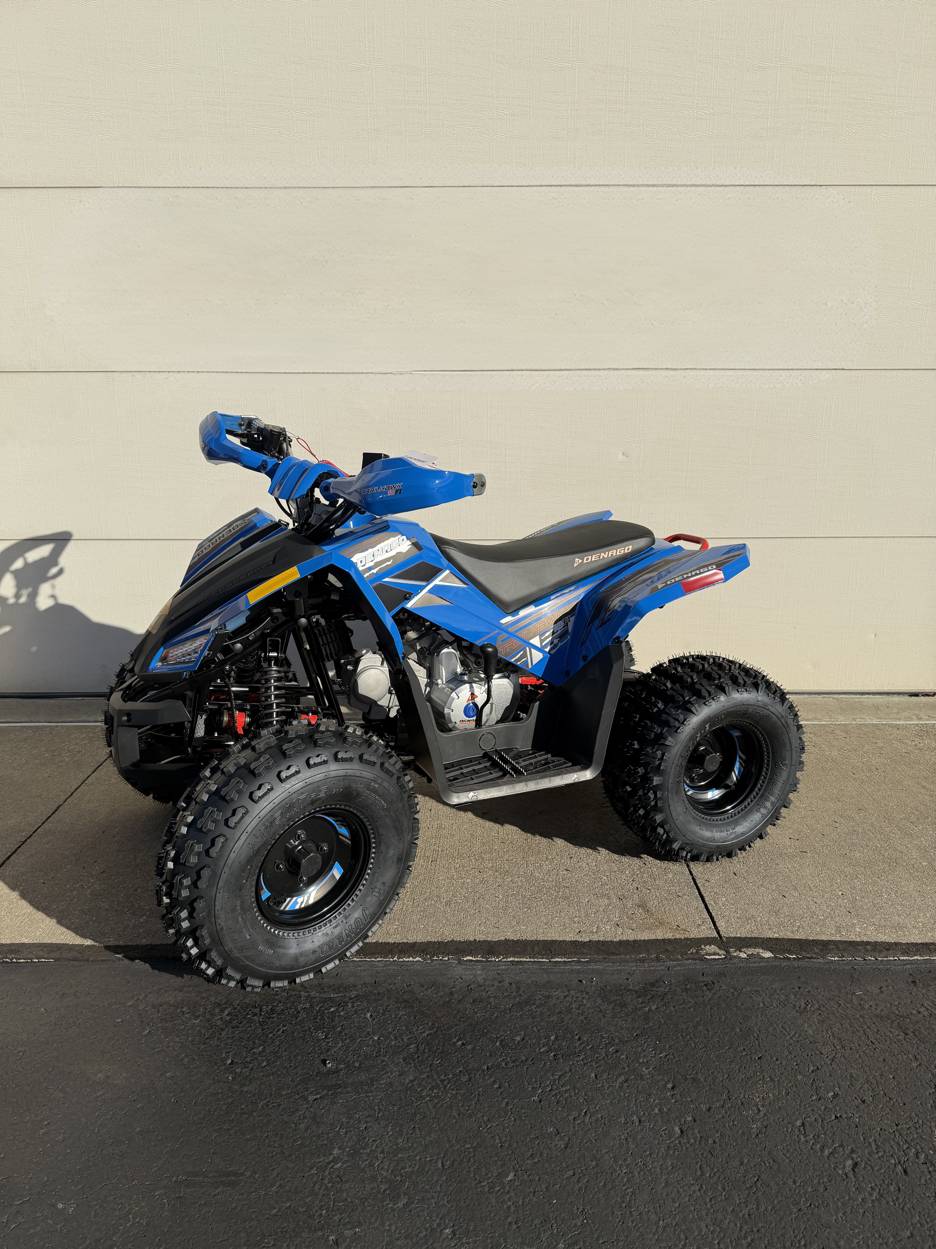 A blue youth-sized ATV with black tires parked on pavement against a beige wall.