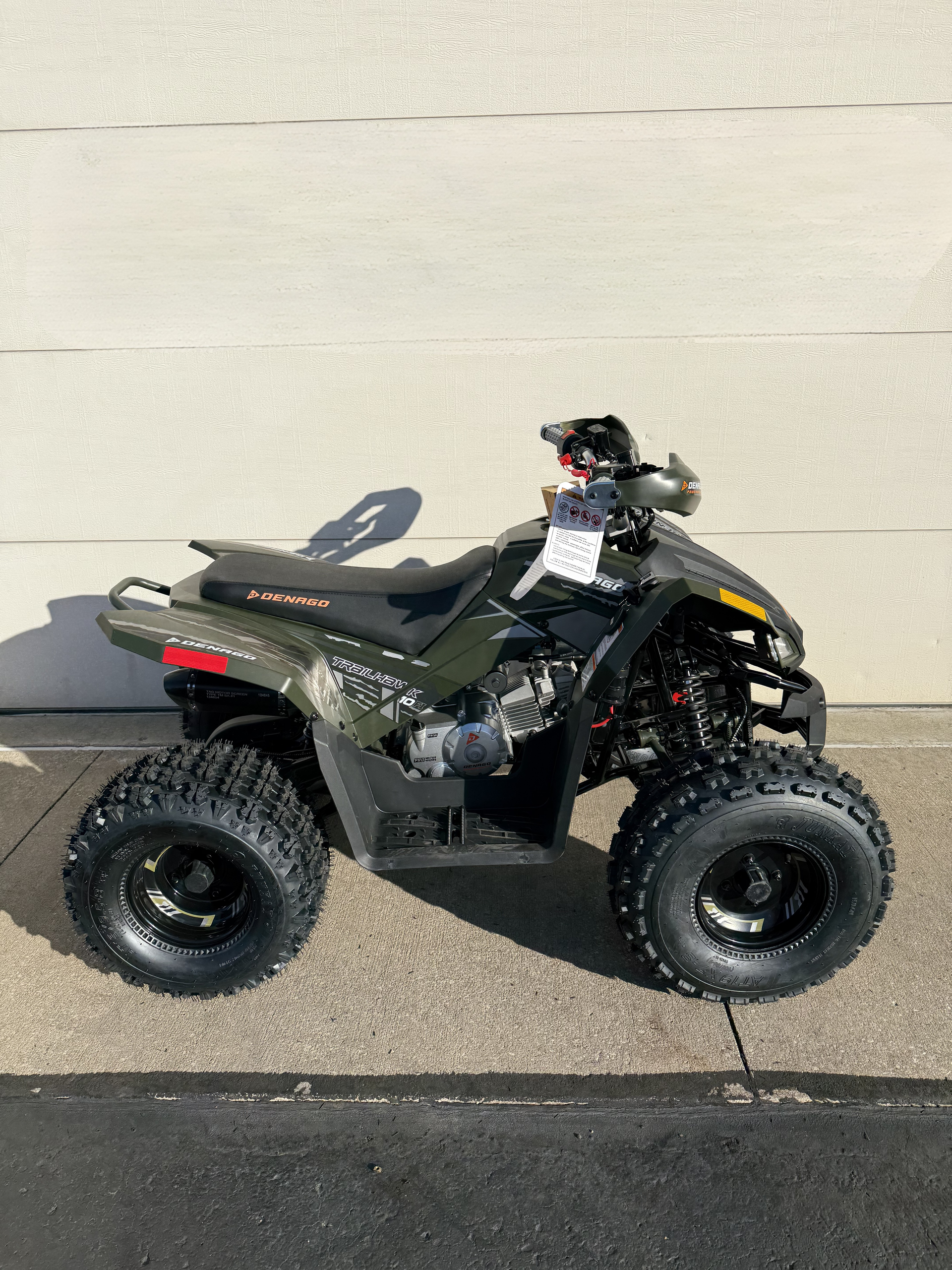 A dark olive green all-terrain vehicle (ATV) with black tires parked on asphalt against a light-colored wall.