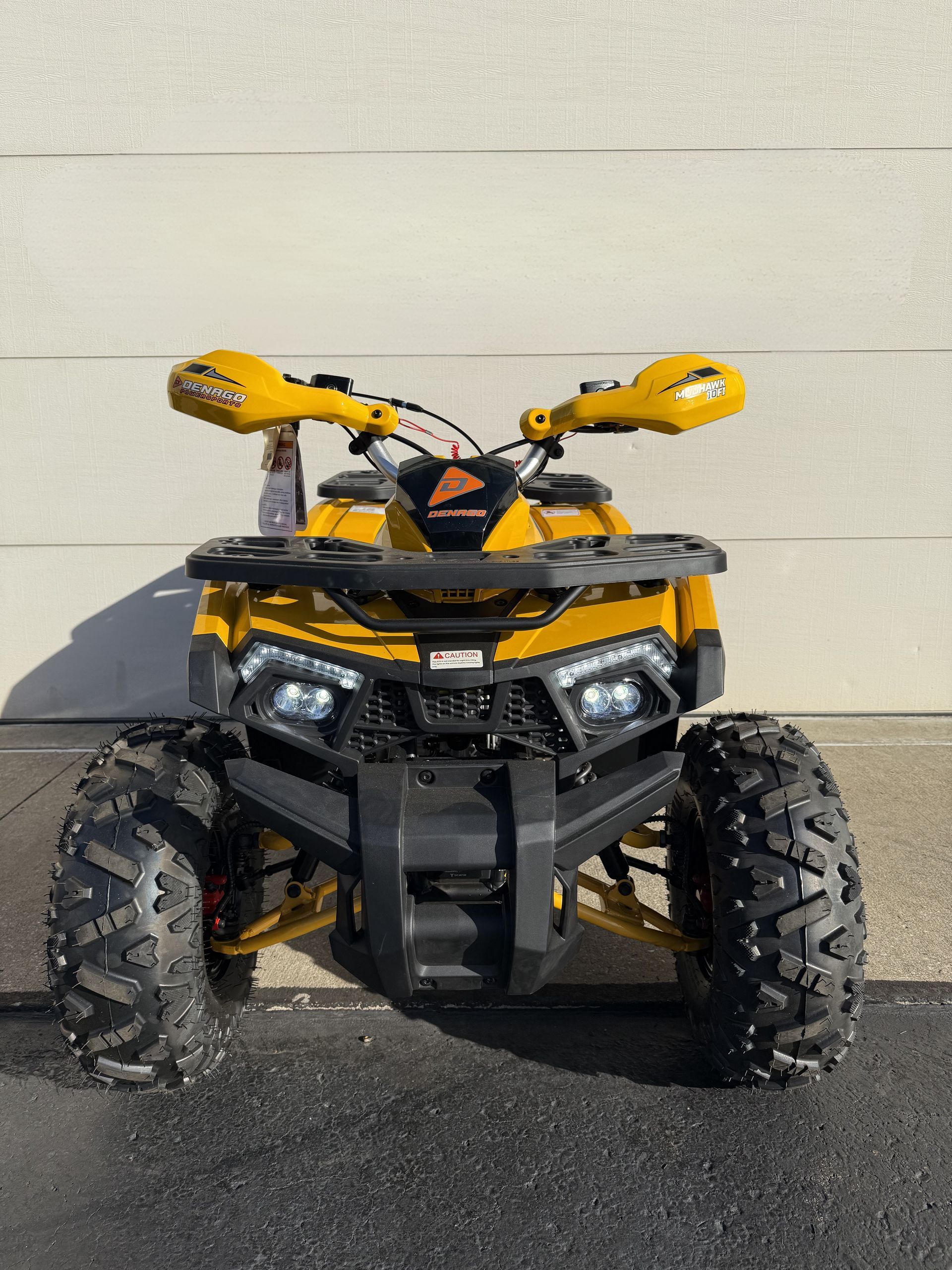 A yellow all-terrain vehicle (ATV) facing forward, parked on pavement against a white wall.