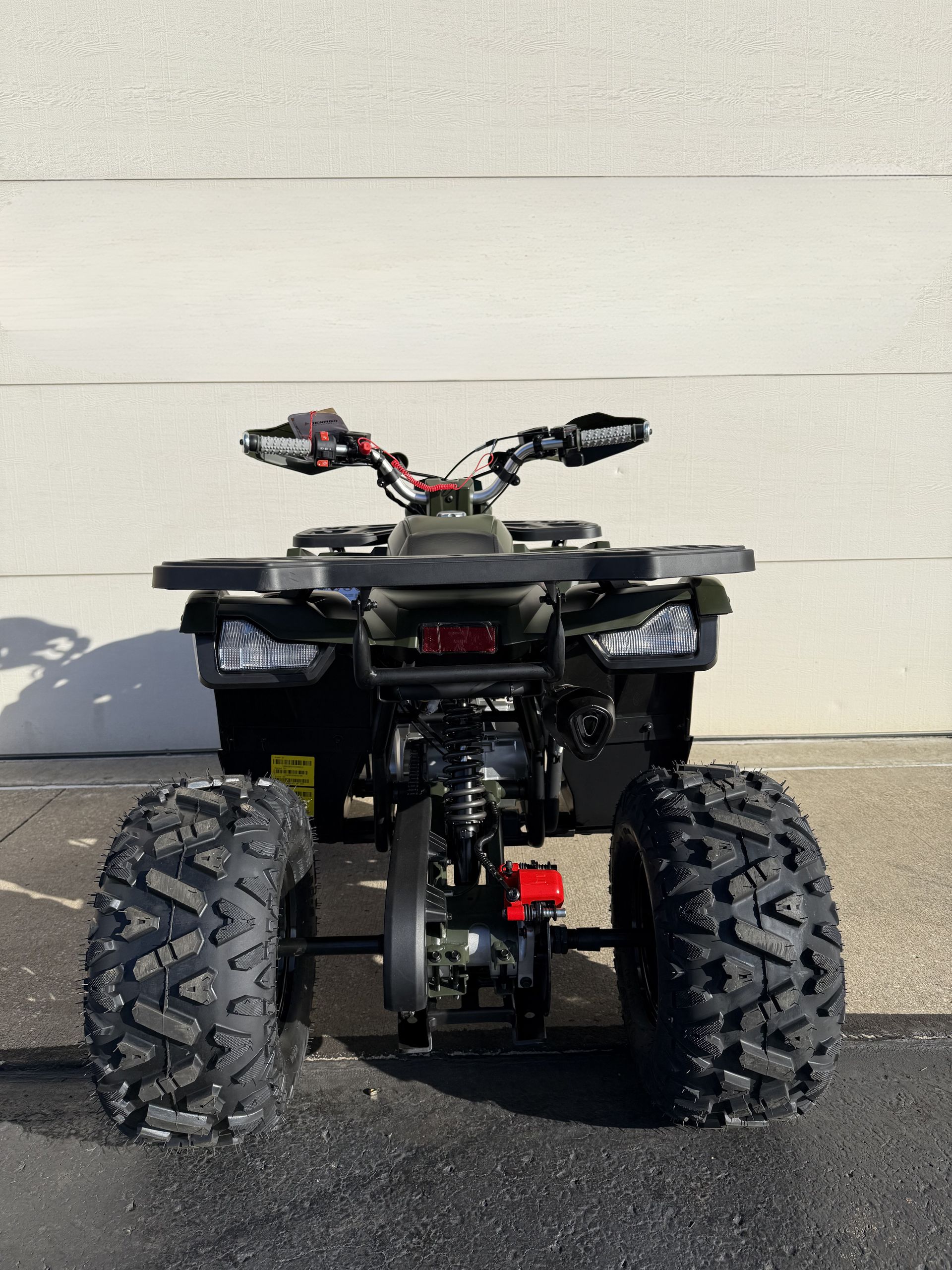 A rear-facing view of a dark-colored ATV with heavy-tread tires parked on asphalt in front of a white wall.