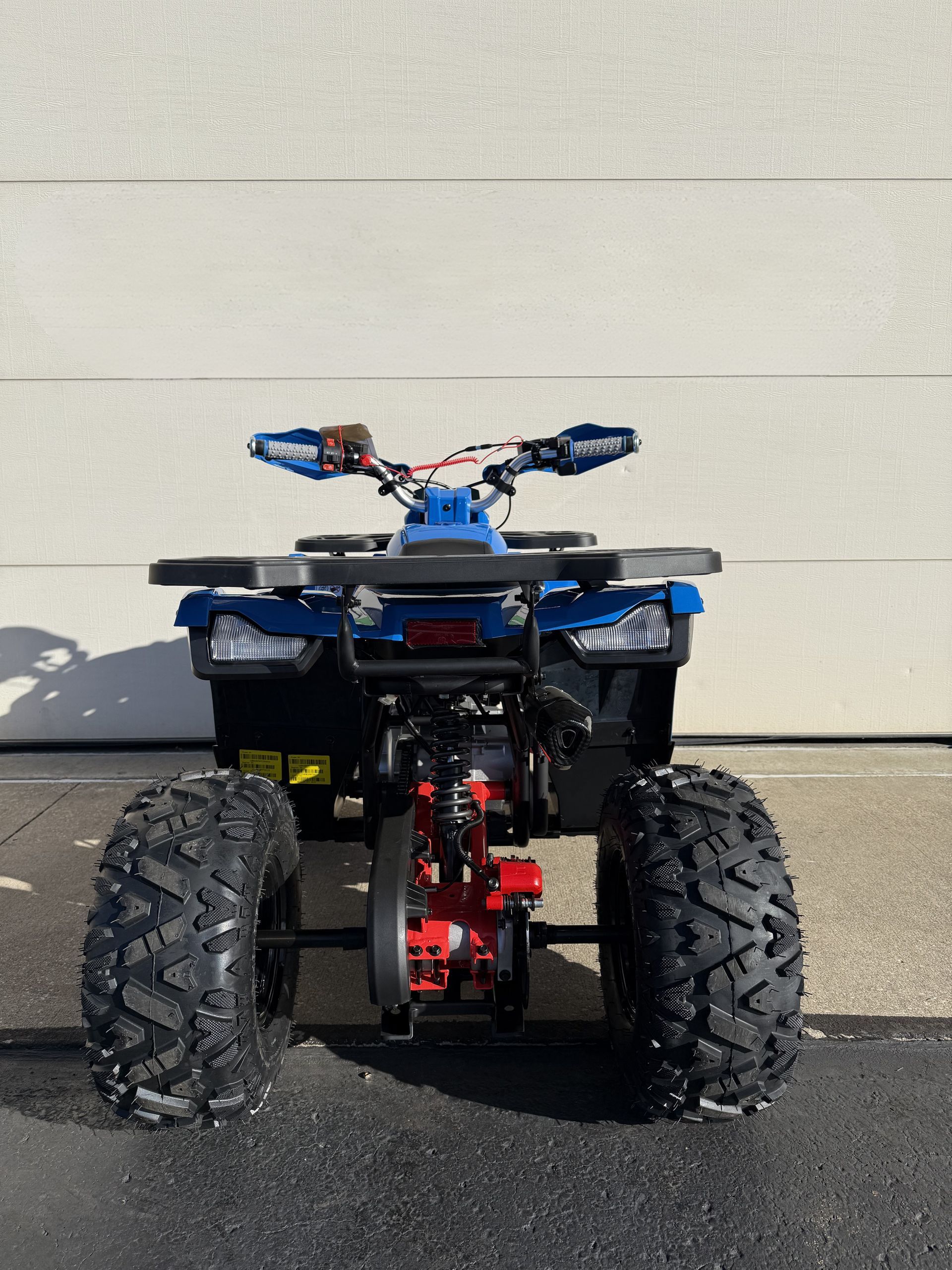 A blue all-terrain vehicle parked on asphalt in front of a white wall, viewed from the rear.
