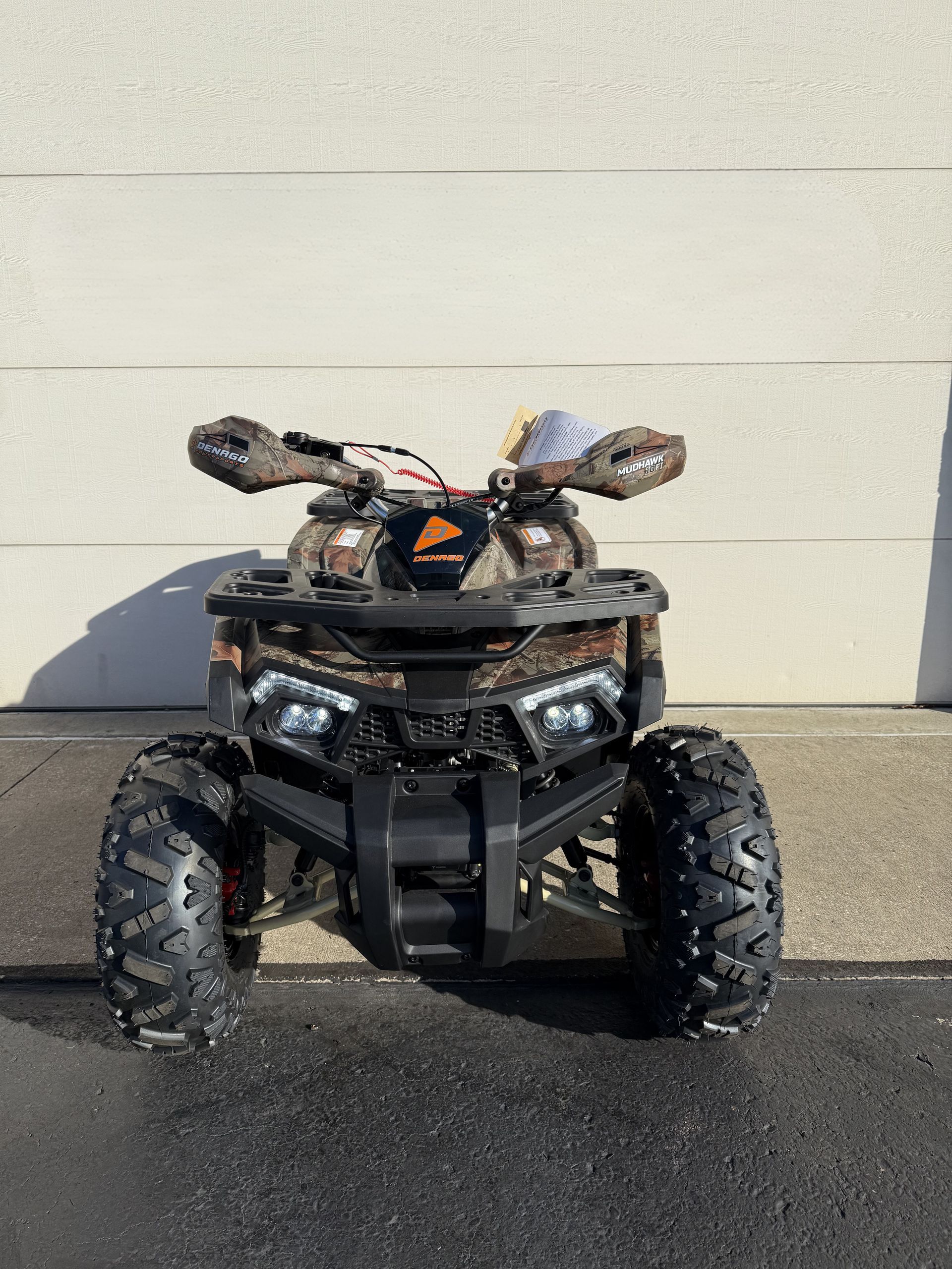 Front view of a camouflage-patterned ATV with black wheels and handguards parked against a beige wall.