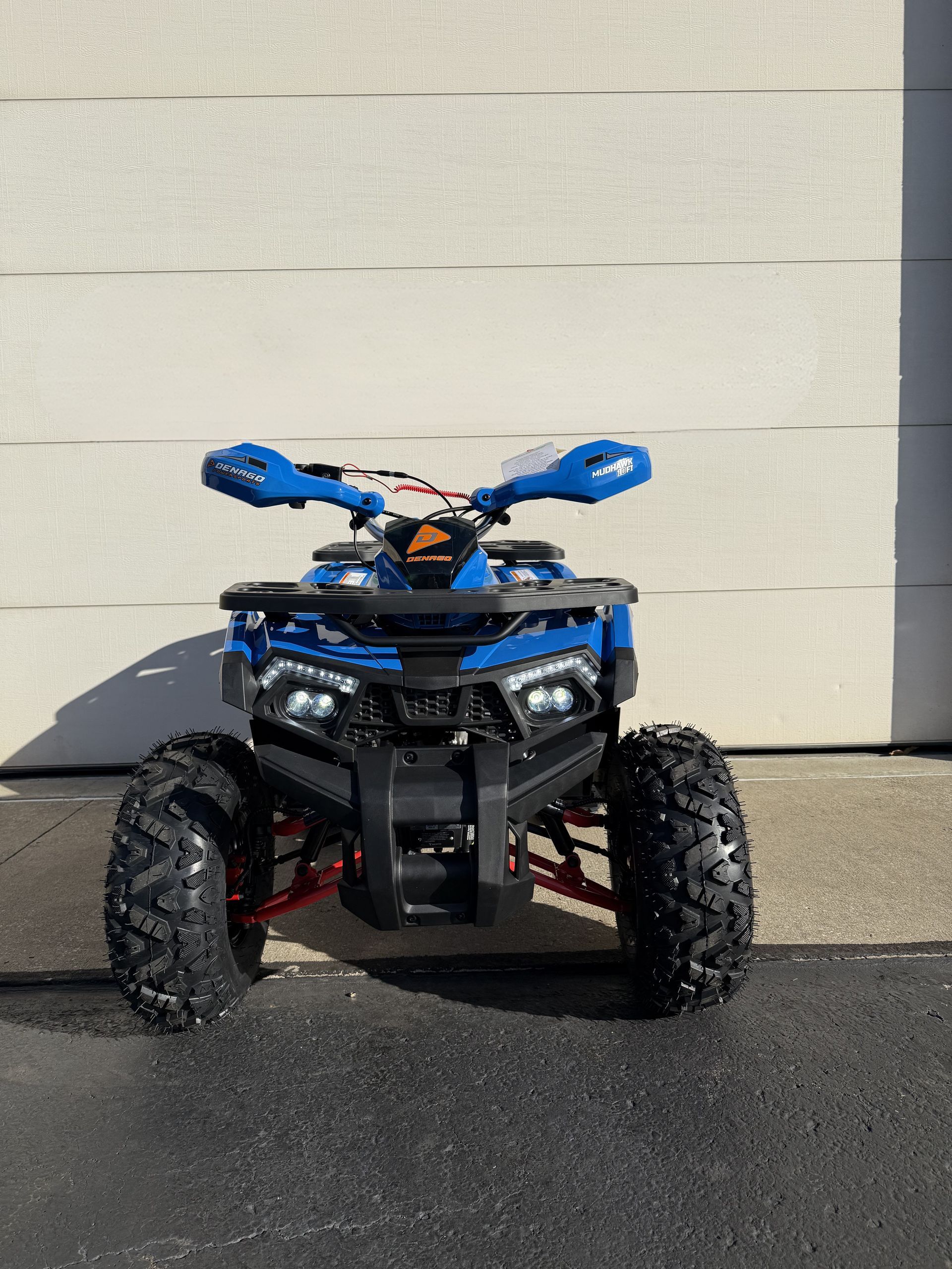 A blue all-terrain vehicle (ATV) with red suspension components parked on pavement in front of a white wall.