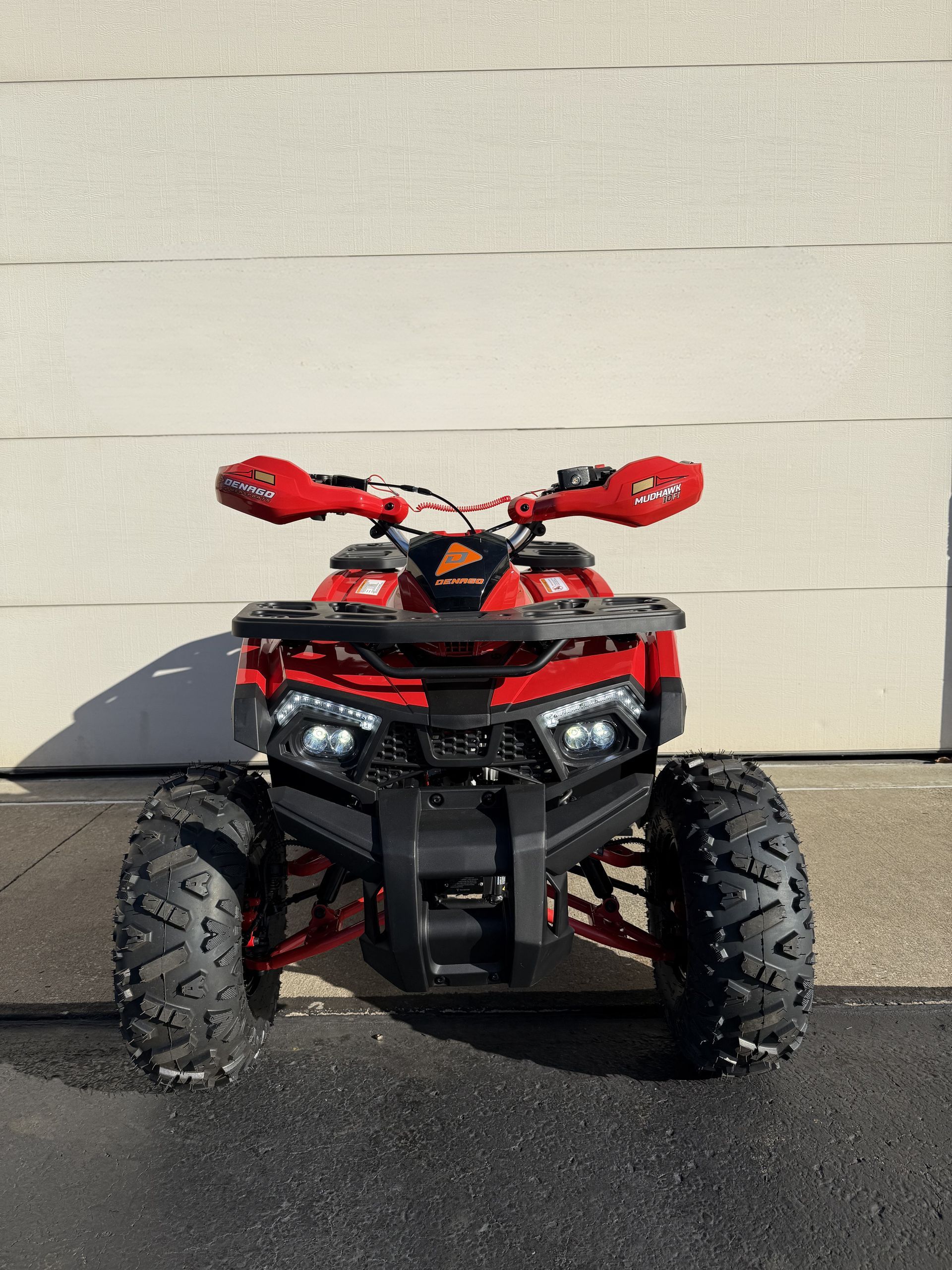 A red ATV parked on pavement in front of a white wall, featuring large black tires and hand guards on the handlebars.