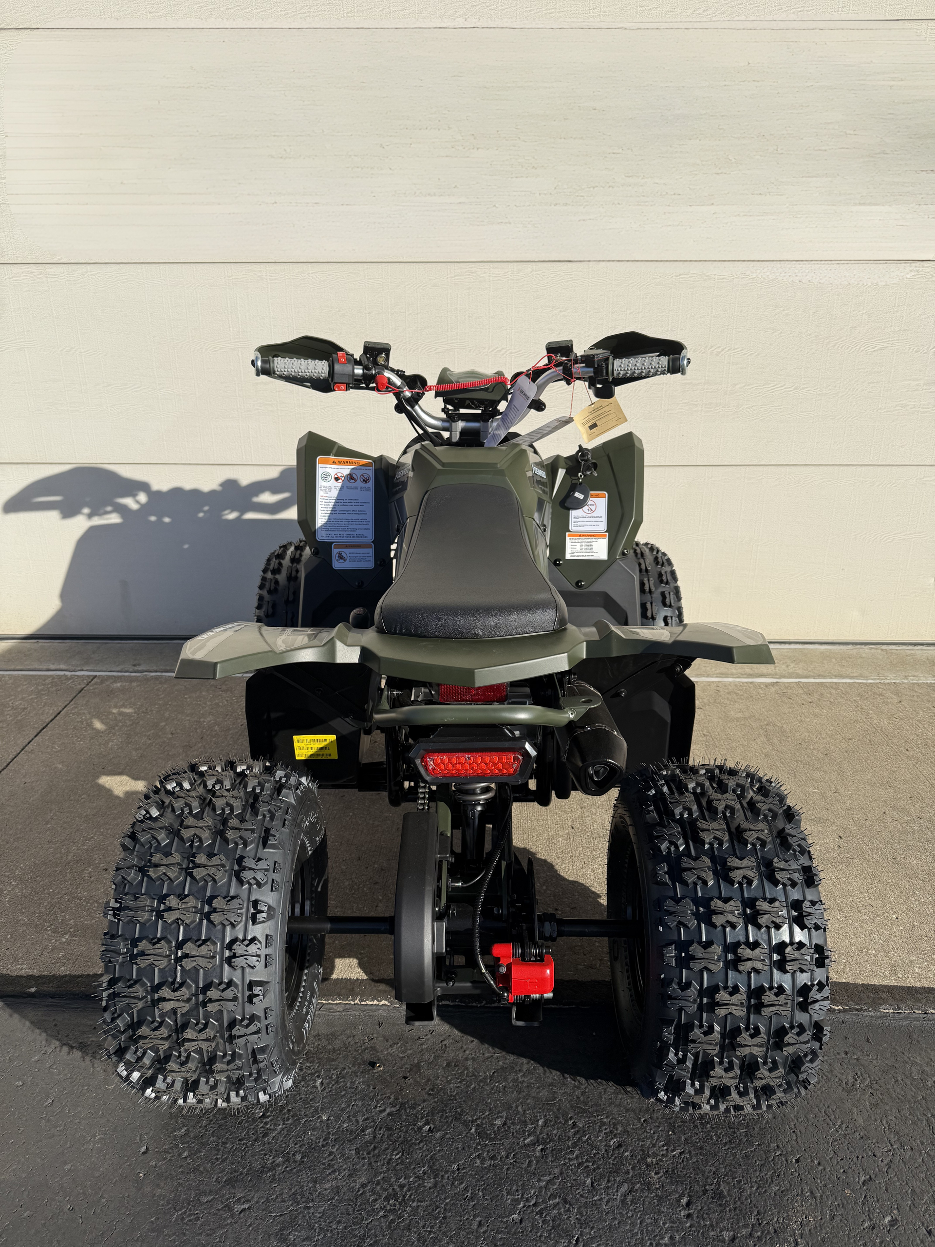 A rear view of an olive green all-terrain vehicle parked on asphalt against a plain wall.