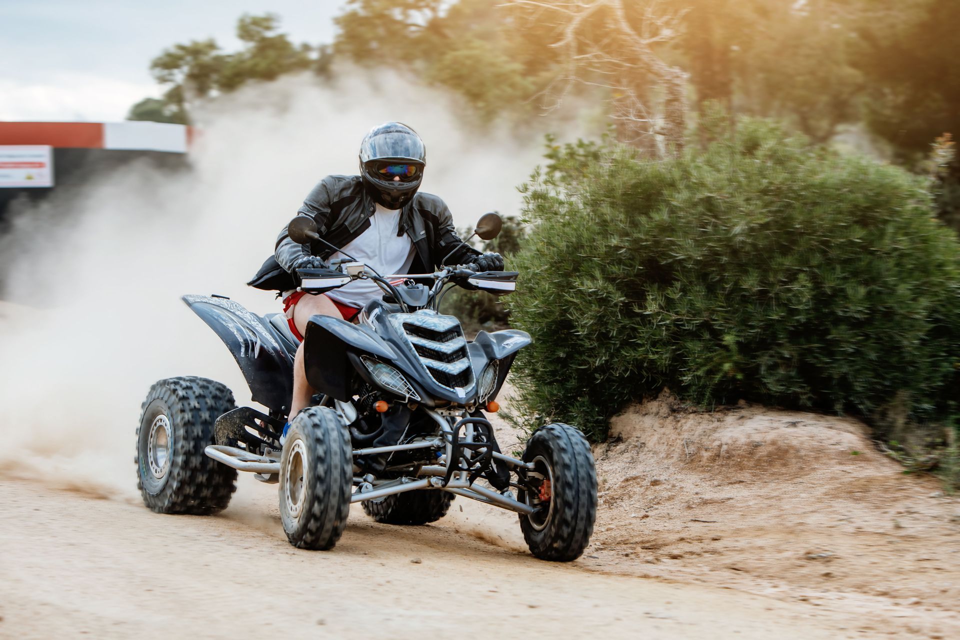 A person in a black leather jacket and helmet riding a black ATV on a dusty dirt path near green bushes.