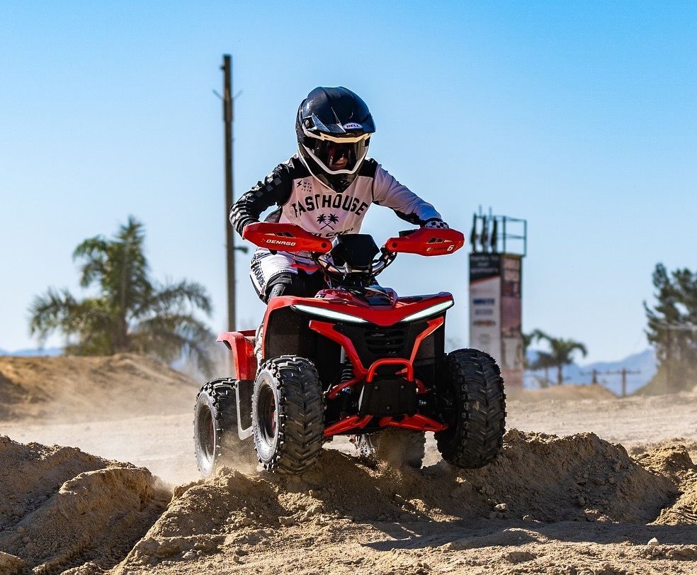 A person in protective gear rides a bright red ATV over a dirt mound on a sunny day.