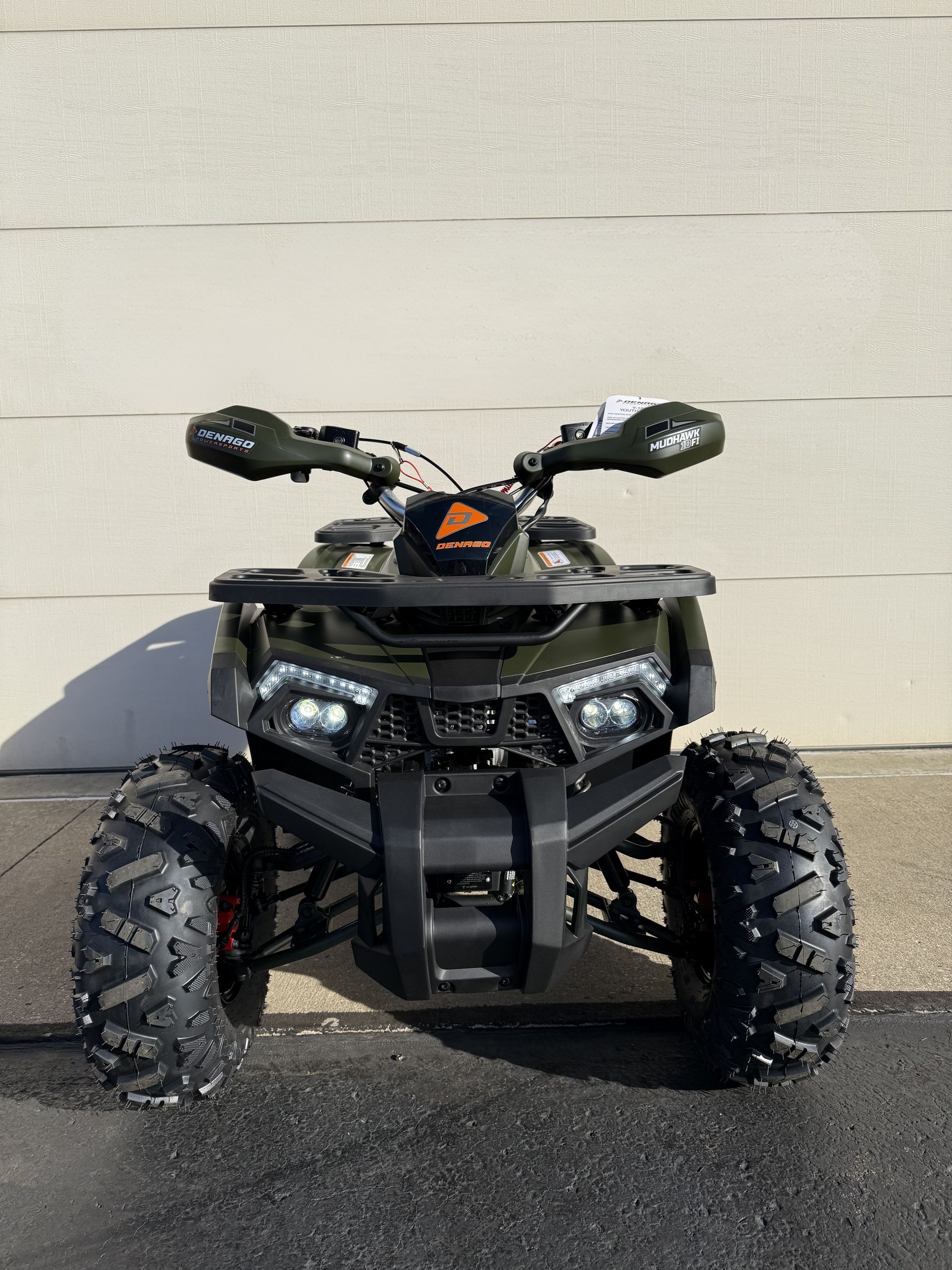 A front-facing, olive green all-terrain vehicle parked on asphalt against a light-colored wall.