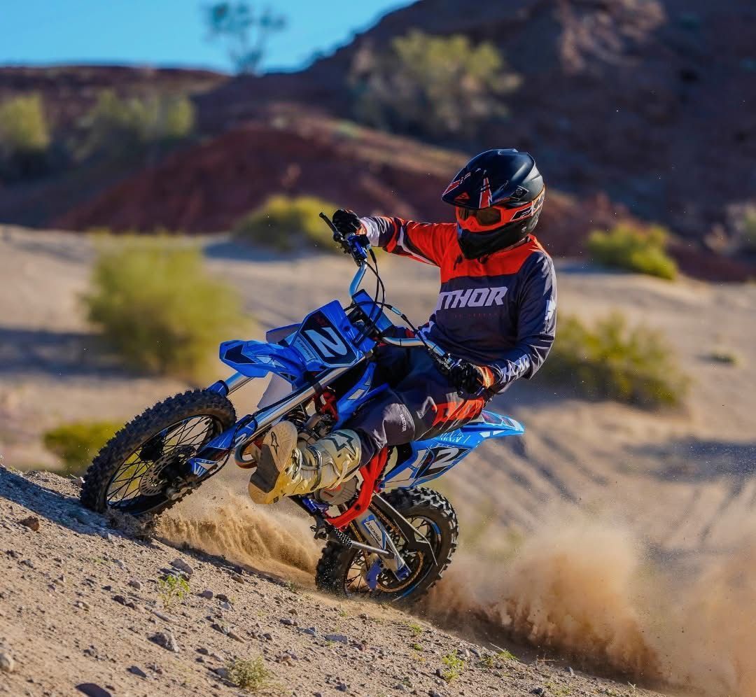 A person in motocross gear rides a blue dirt bike while turning on a dusty, sandy hill.