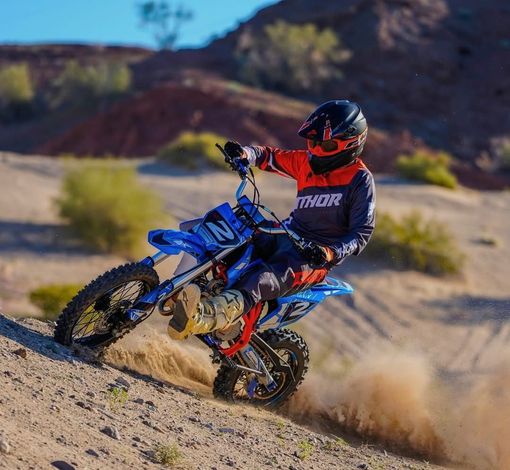 A person in motocross gear rides a blue dirt bike while turning on a dusty, sandy hill.
