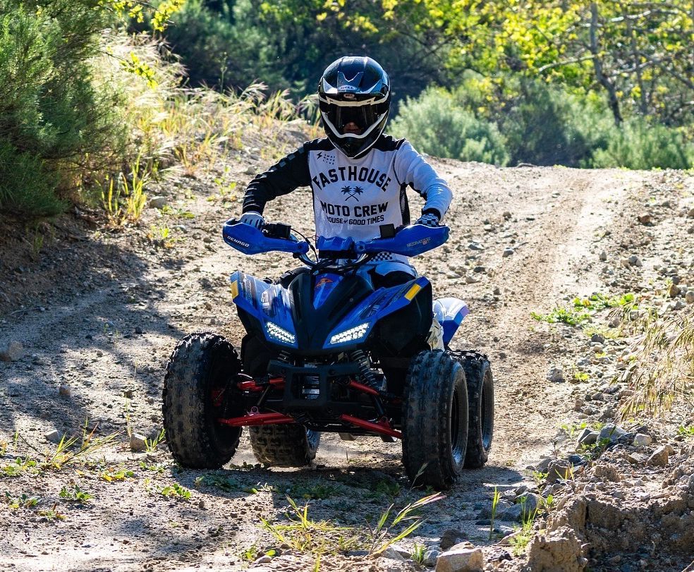 A person in protective gear rides a blue all-terrain vehicle on a dirt trail in a sunlit forest.