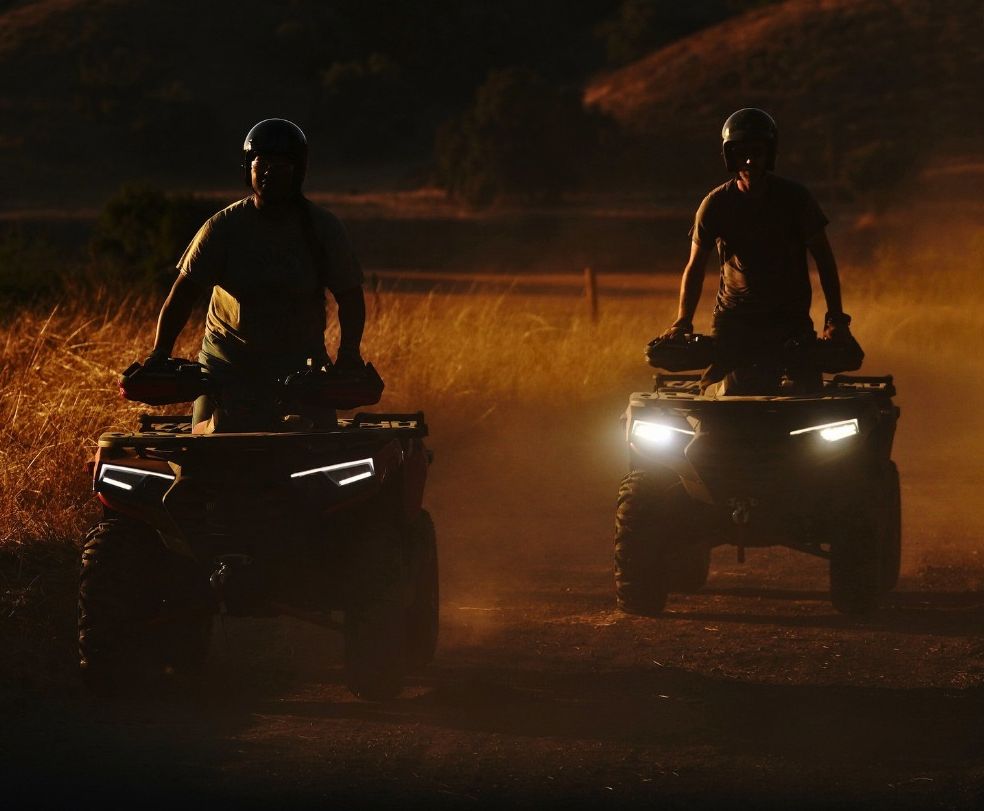 Two people riding all-terrain vehicles through a dusty, sunlit field at dusk.