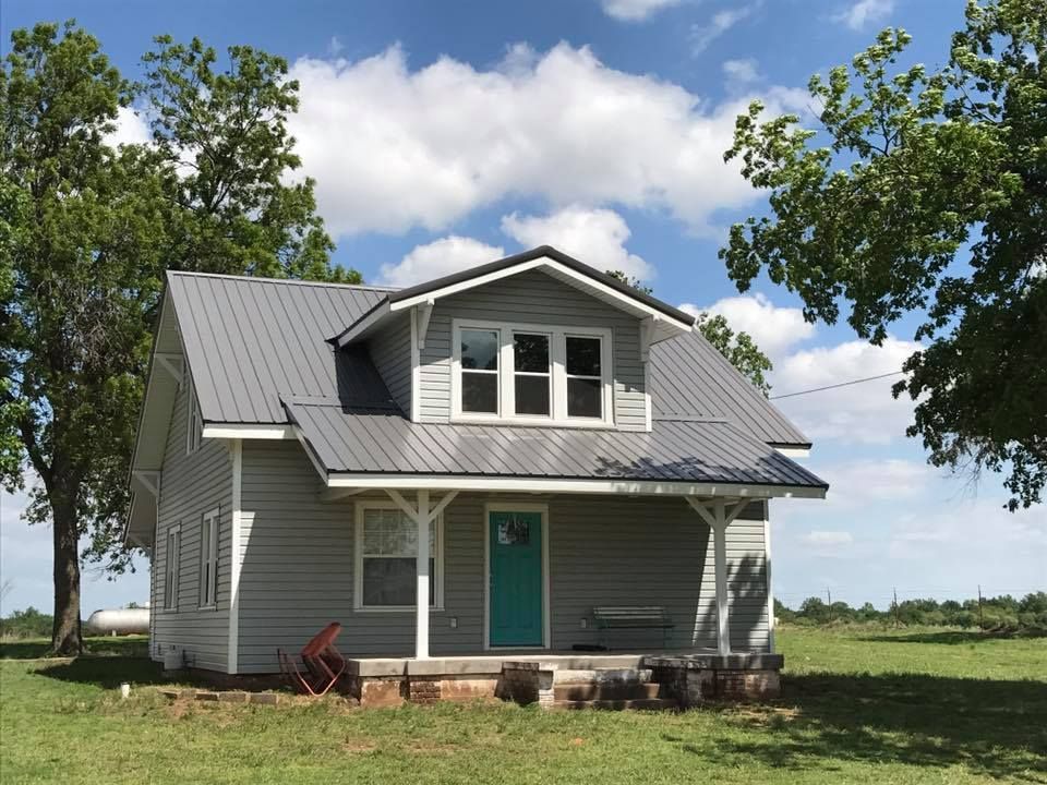 Small gray house with teal door and metal roof. Trees and grass surround it.