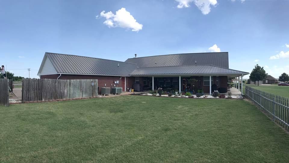 Backyard view of a brick house with a metal roof, green lawn, wooden fence, and blue sky with clouds.