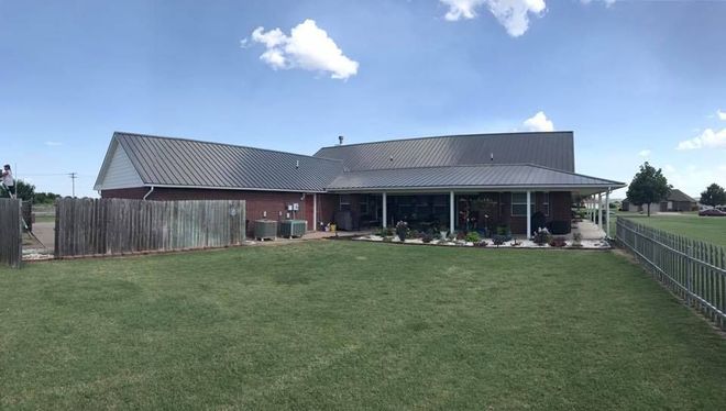Backyard view of a brick house with a metal roof, green lawn, wooden fence, and blue sky with clouds.