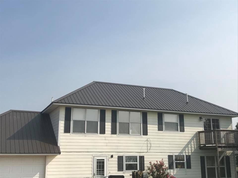 Two-story house with dark gray metal roof, white siding, black shutters, and wooden deck under blue sky.