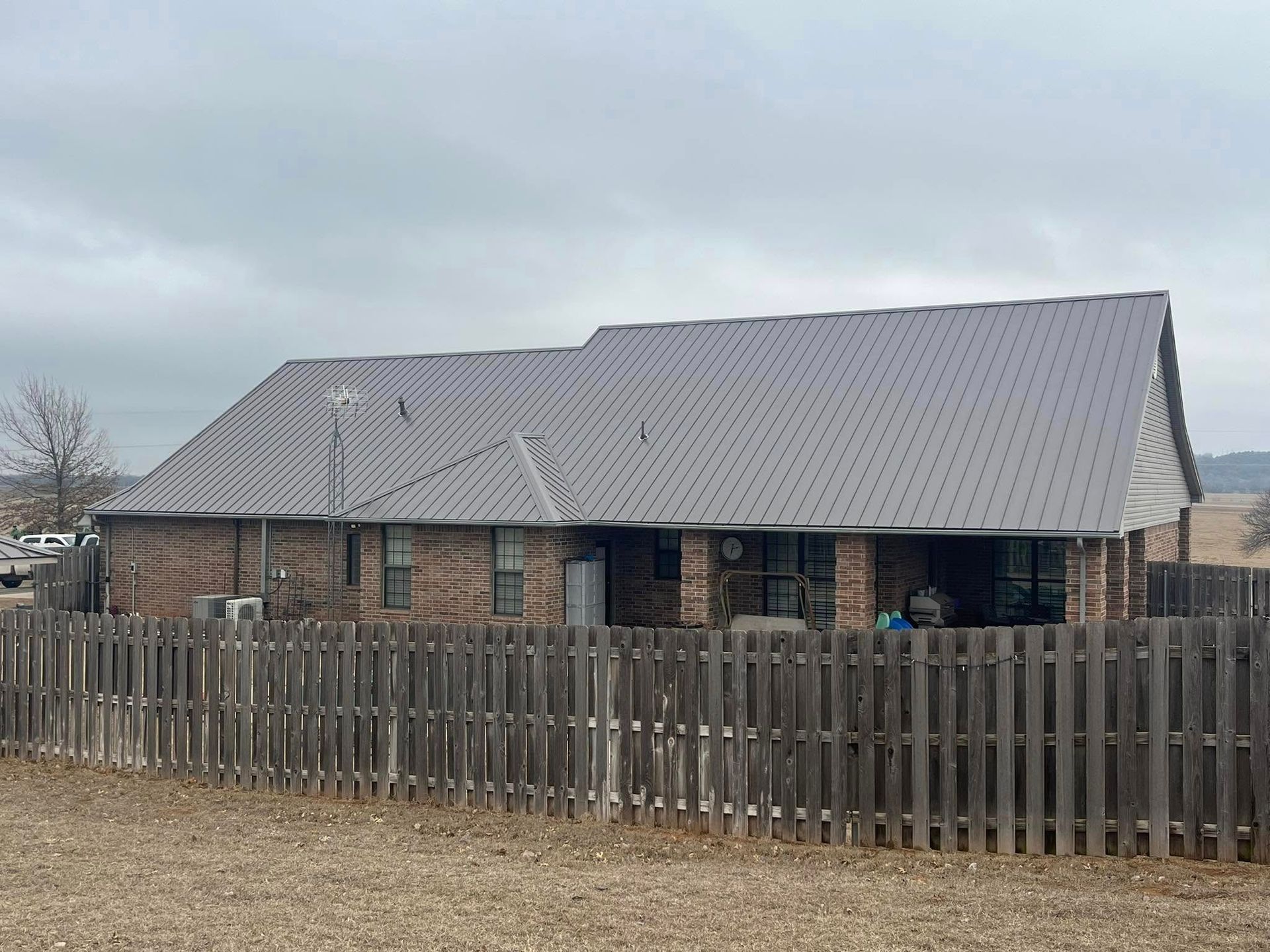 Brown brick house with gray metal roof behind a wooden fence under a cloudy sky.