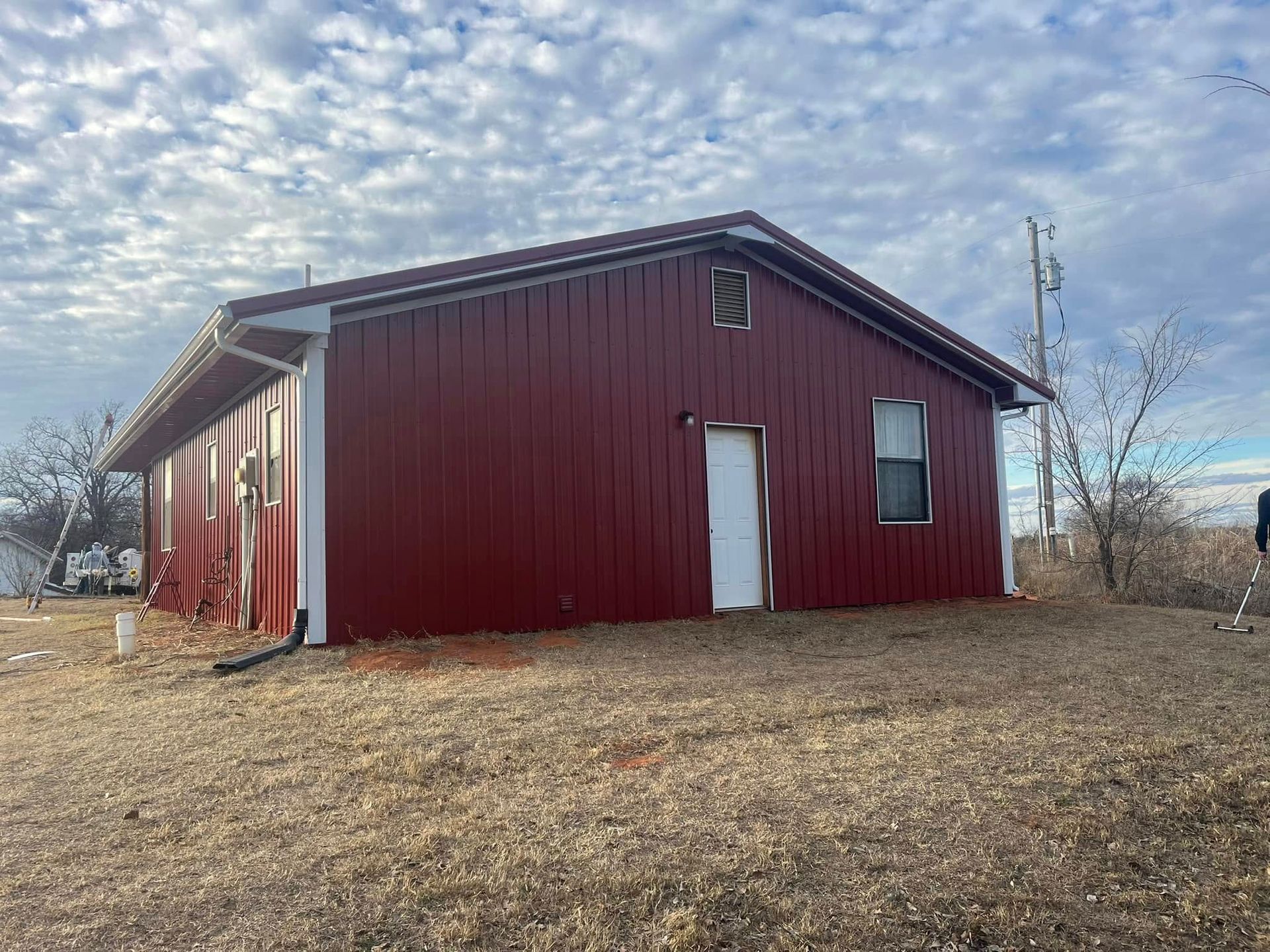 Red metal-clad building with a white door and window on a grassy field, under a cloudy sky.