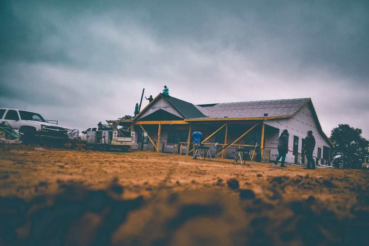 Construction of a house under cloudy sky. People working on a partially built building; a truck is parked nearby.